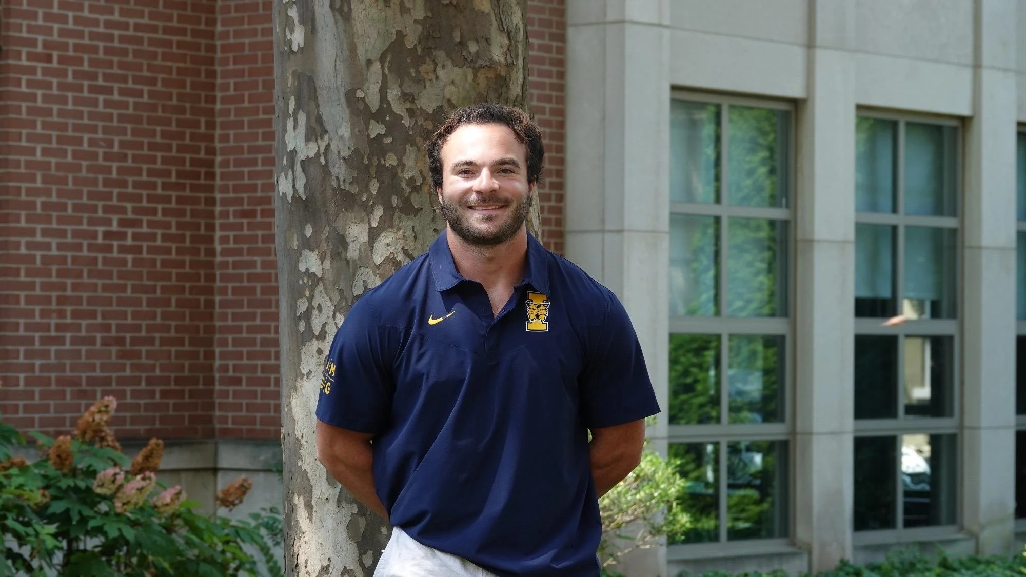 A young man with dark hair and a beard, smiling, wearing a navy blue polo shirt with a yellow logo, standing outdoors next to a tree with a brick building and windows in the background.