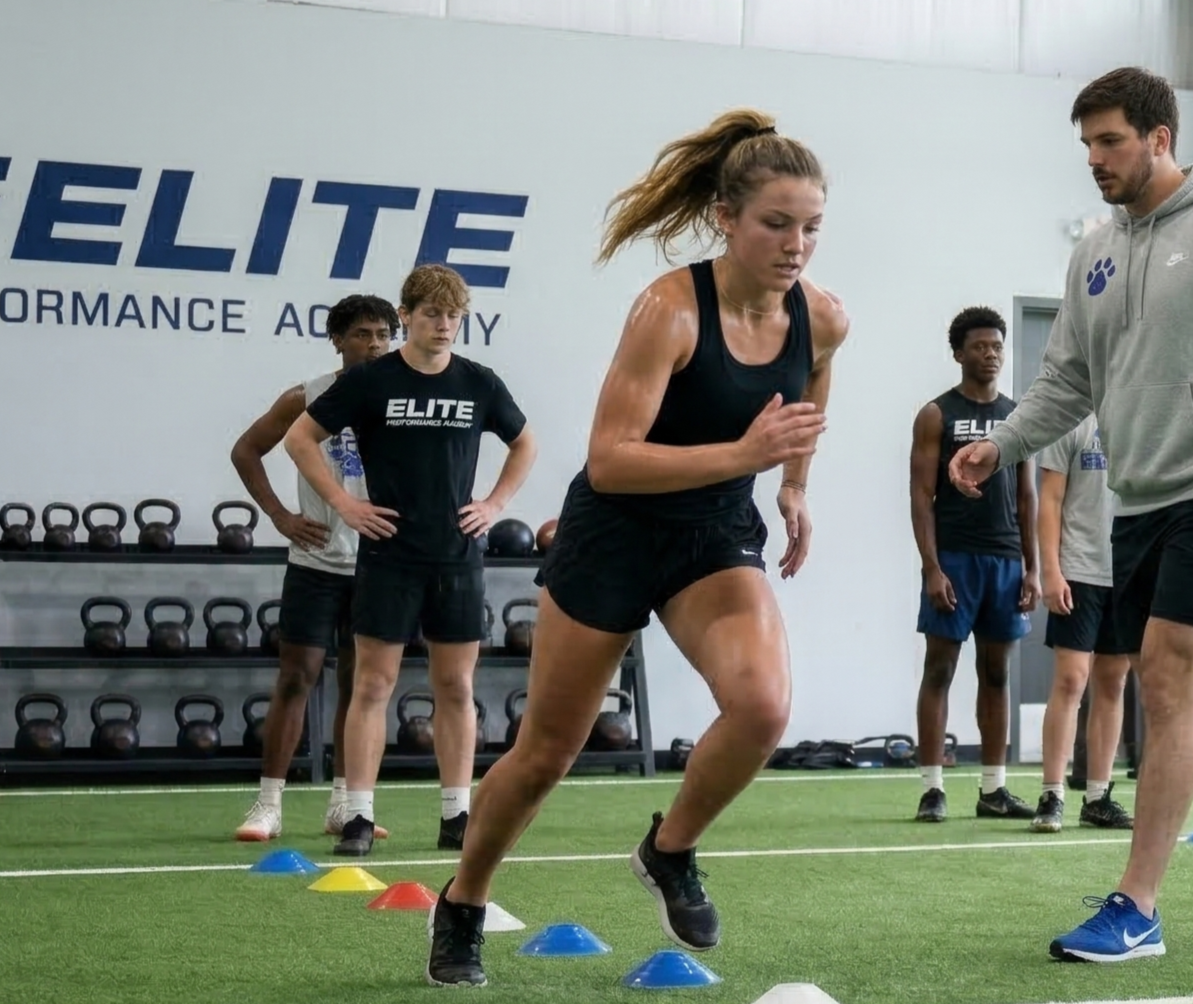 Young woman running through agility cones in a gym with a coach and teammates watching.
