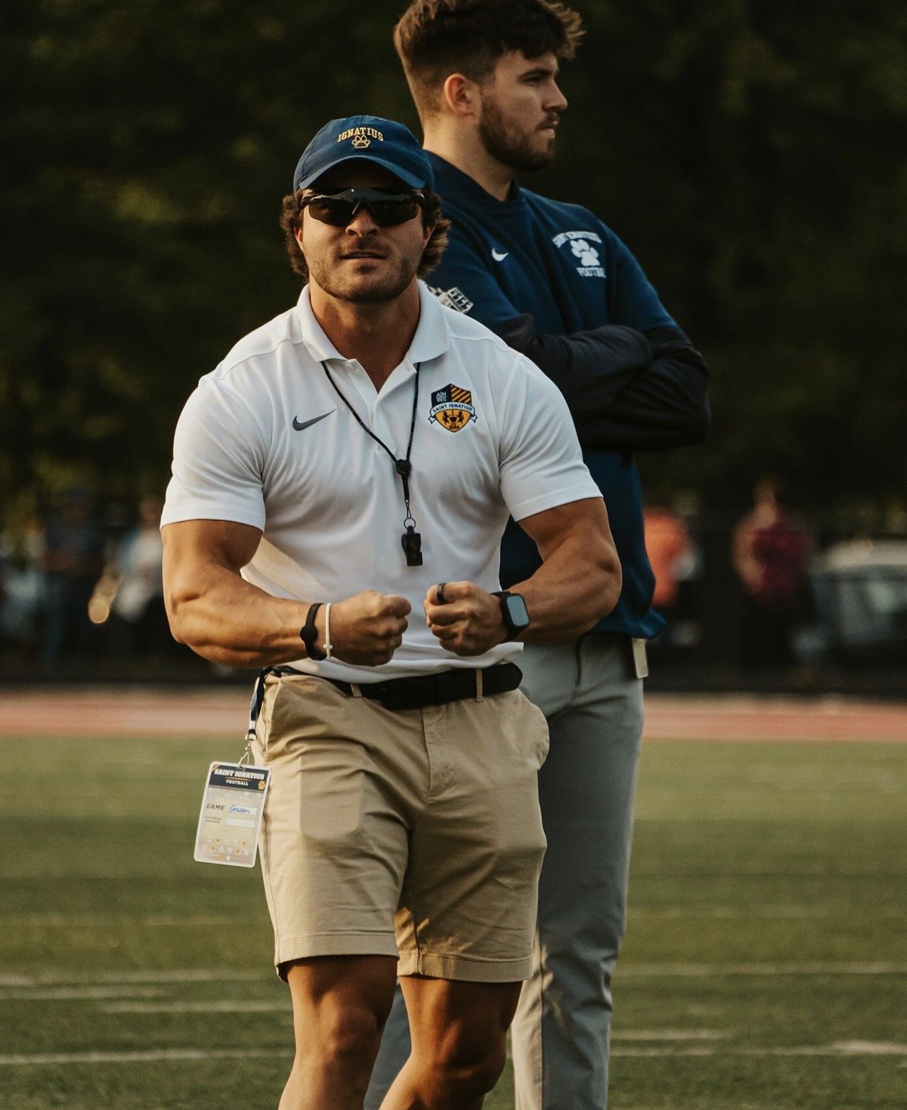 A man with a muscular build wearing sunglasses, a white polo shirt, beige shorts, and a hat with a whistle around his neck, standing on a sports field with a man in a blue jacket behind him.