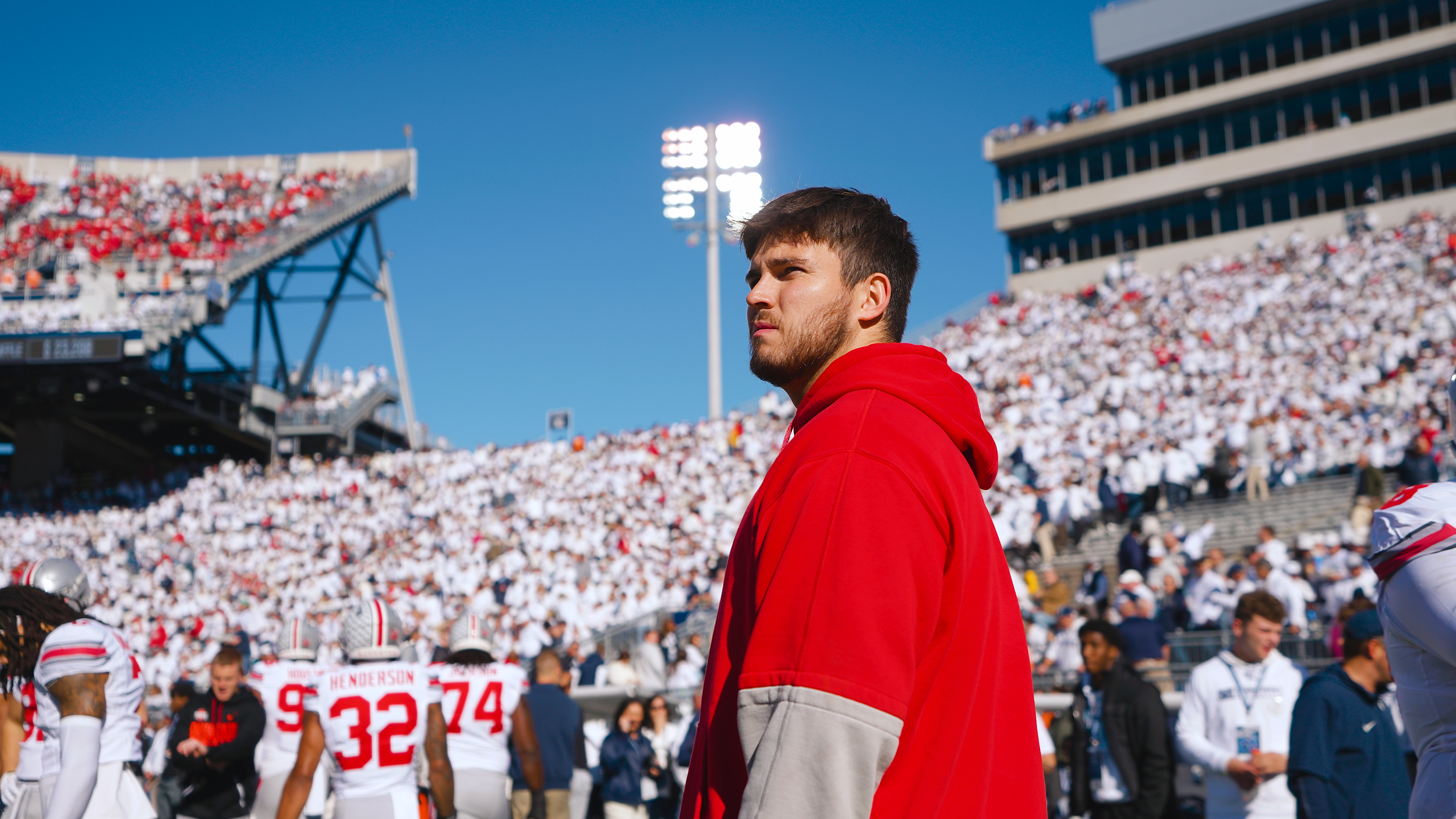 A football player in a red hoodie stands on the sideline of a stadium filled with spectators, with bright stadium lights and a clear blue sky overhead.