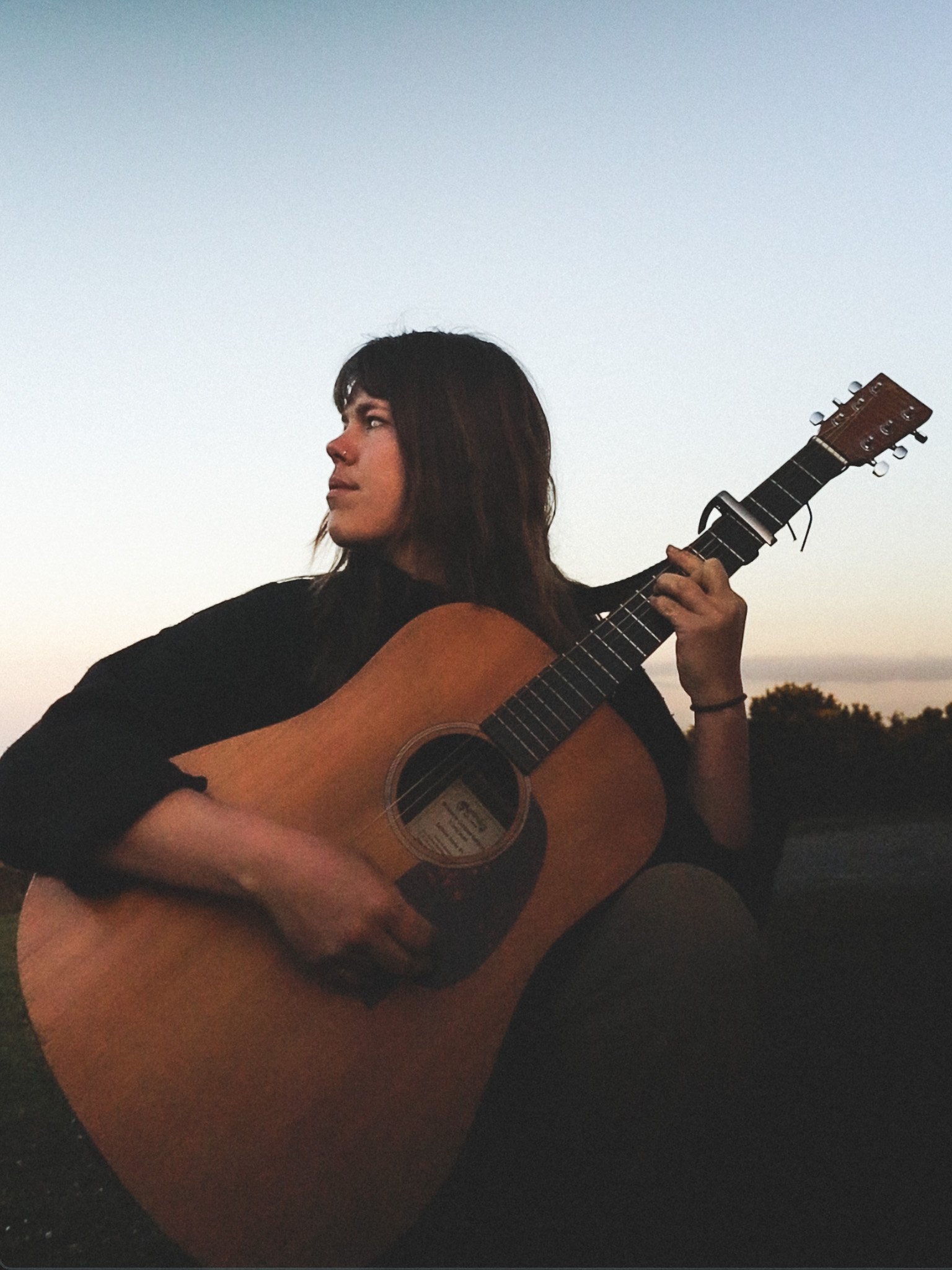 A woman with dark hair is sitting outdoors at dusk, playing an acoustic guitar with sheet music inside the sound hole, with a background of trees and a fading sky.