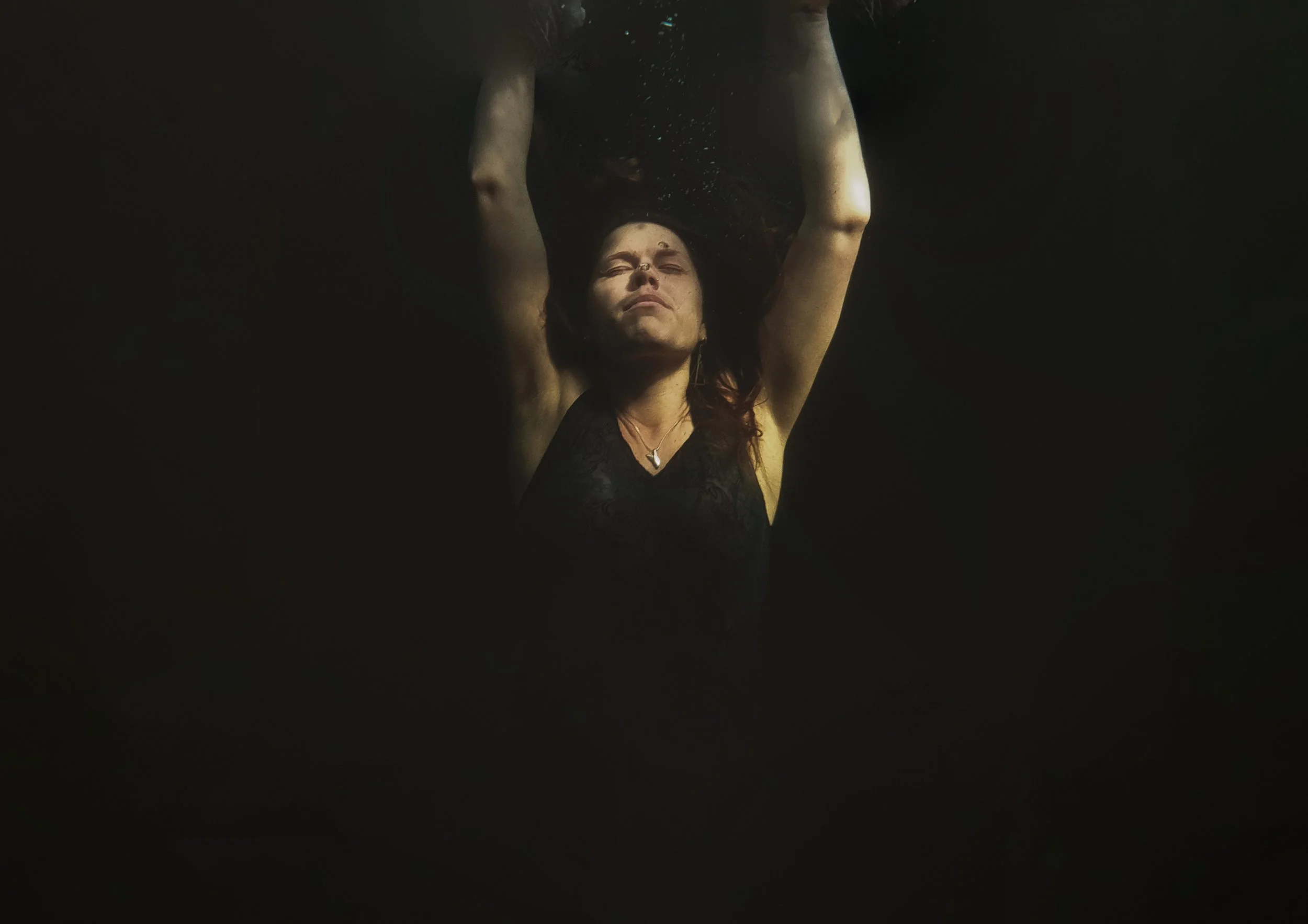 A woman is underwater with her arms raised above her head, wearing a black top and a necklace, with her eyes closed and face relaxed.