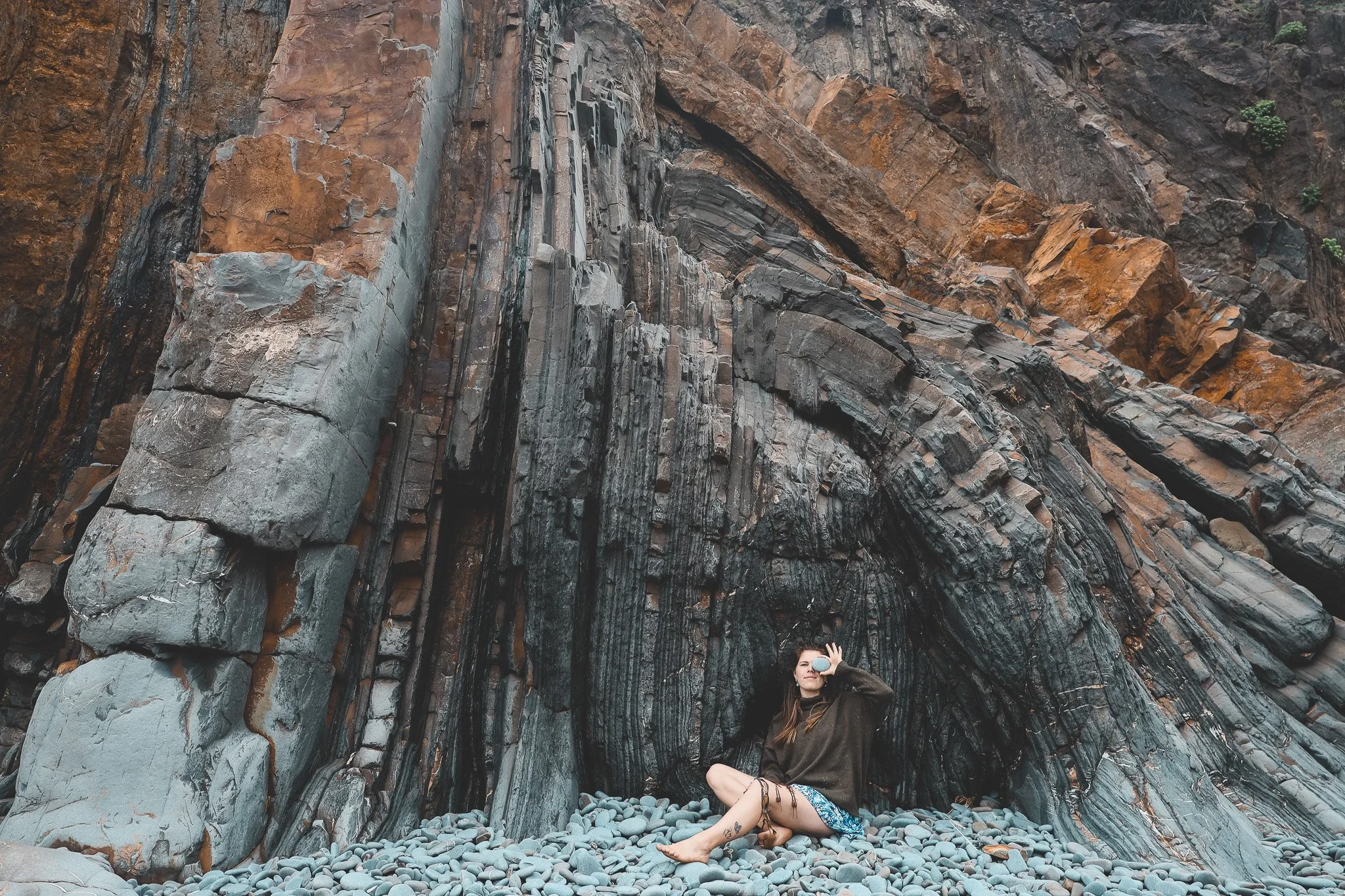 A woman sitting cross-legged with her eyes closed in front of a large, rocky cliff. She is wearing a dark jacket and blue patterned shorts, and is holding a round mirror to her face. The base of the cliff has smooth, rounded stones.
