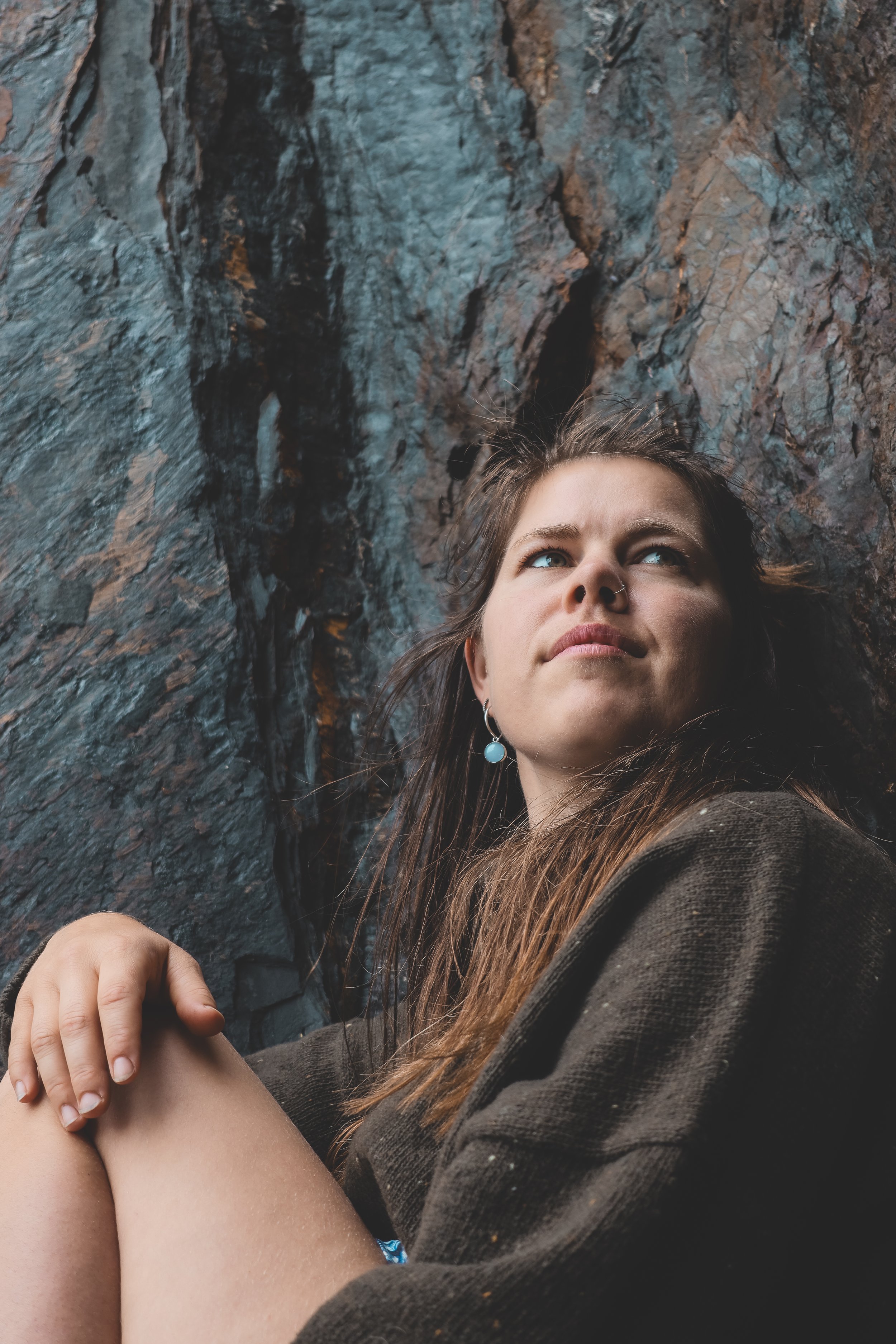 A young woman with long brown hair, wearing earrings, is resting her head against a rocky cliff face, looking upward with a calm expression.