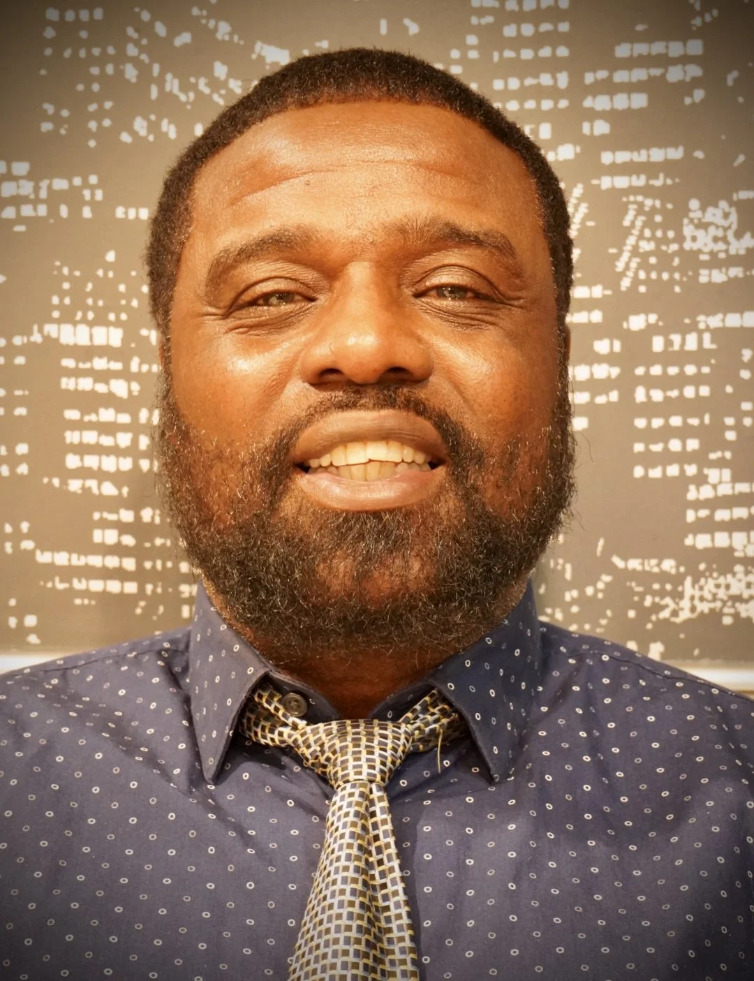 Close-up of a smiling man with a beard, wearing a blue patterned shirt and a tied necktie, with an abstract black and white background.