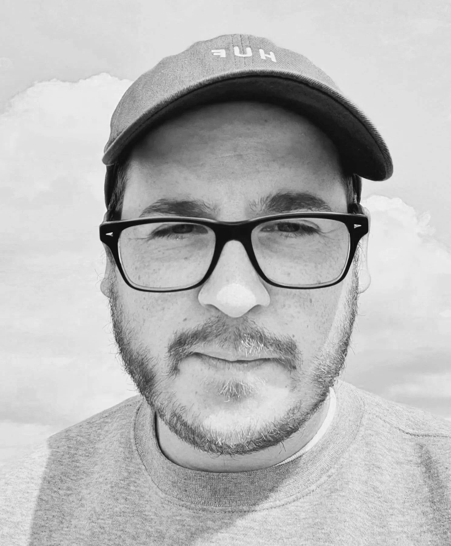 Black and white close-up photo of a man wearing glasses, a cap, and a sweatshirt, outdoors with clouds in the background.