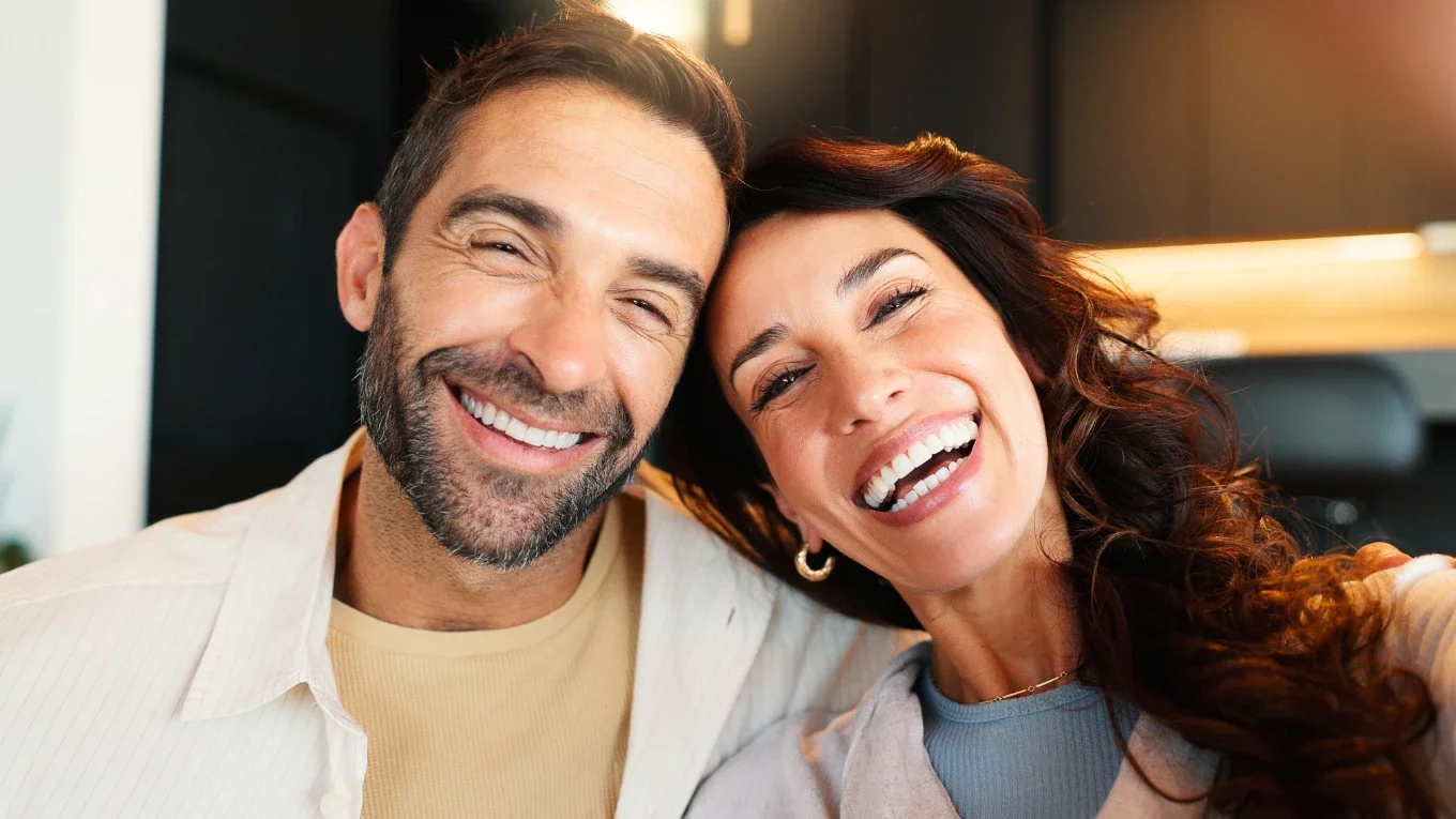 Close-up of two smiling adults, man with short dark hair and beard, woman with long curly brown hair, taking a selfie indoors.