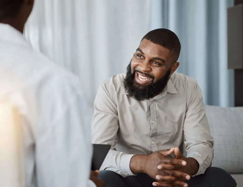 A man with a beard smiling and engaging in conversation with a woman, both seated indoors.