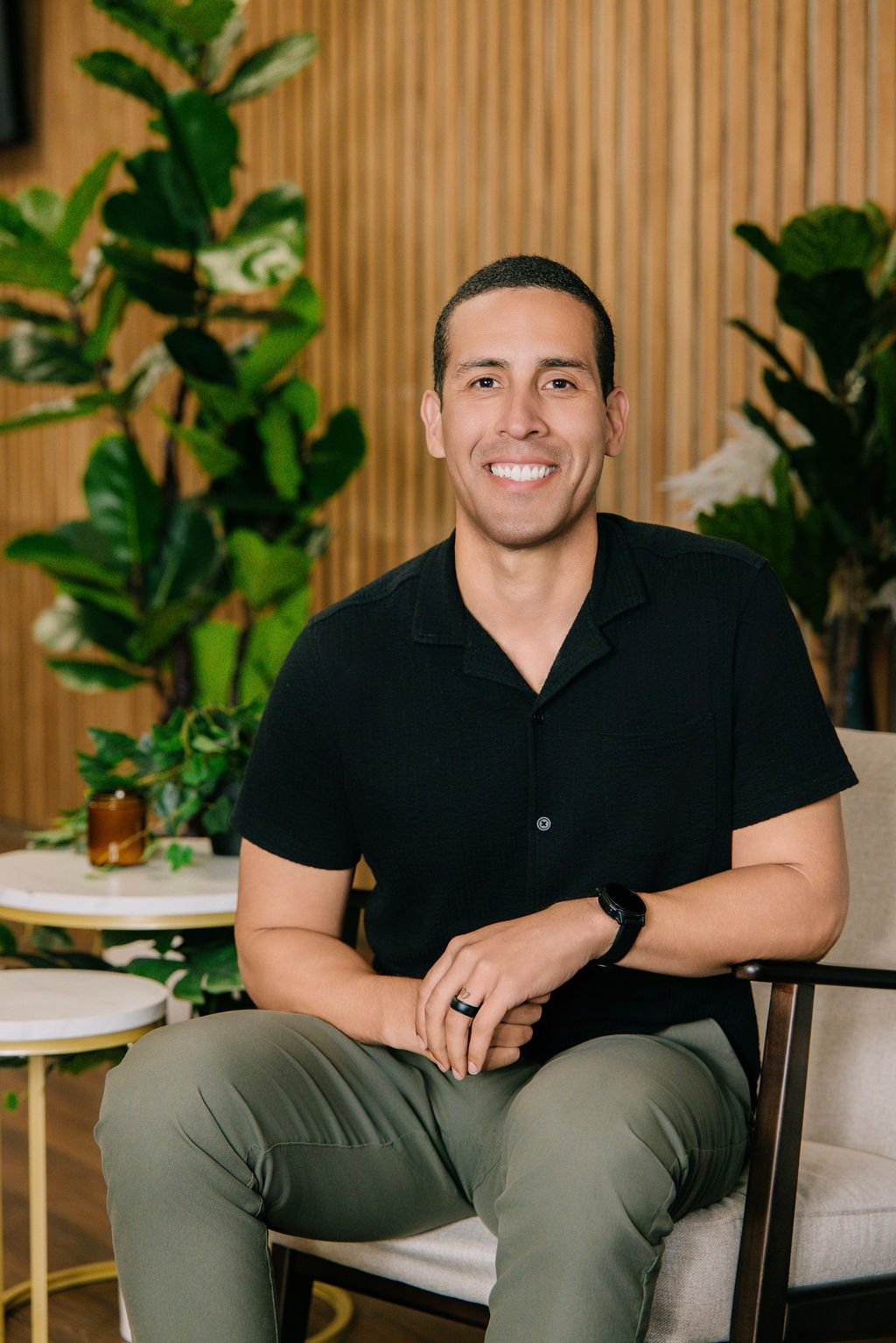A smiling man with short dark hair, wearing a black polo shirt and beige pants, sitting on a chair in a modern indoor setting with wooden wall panels and green plants.