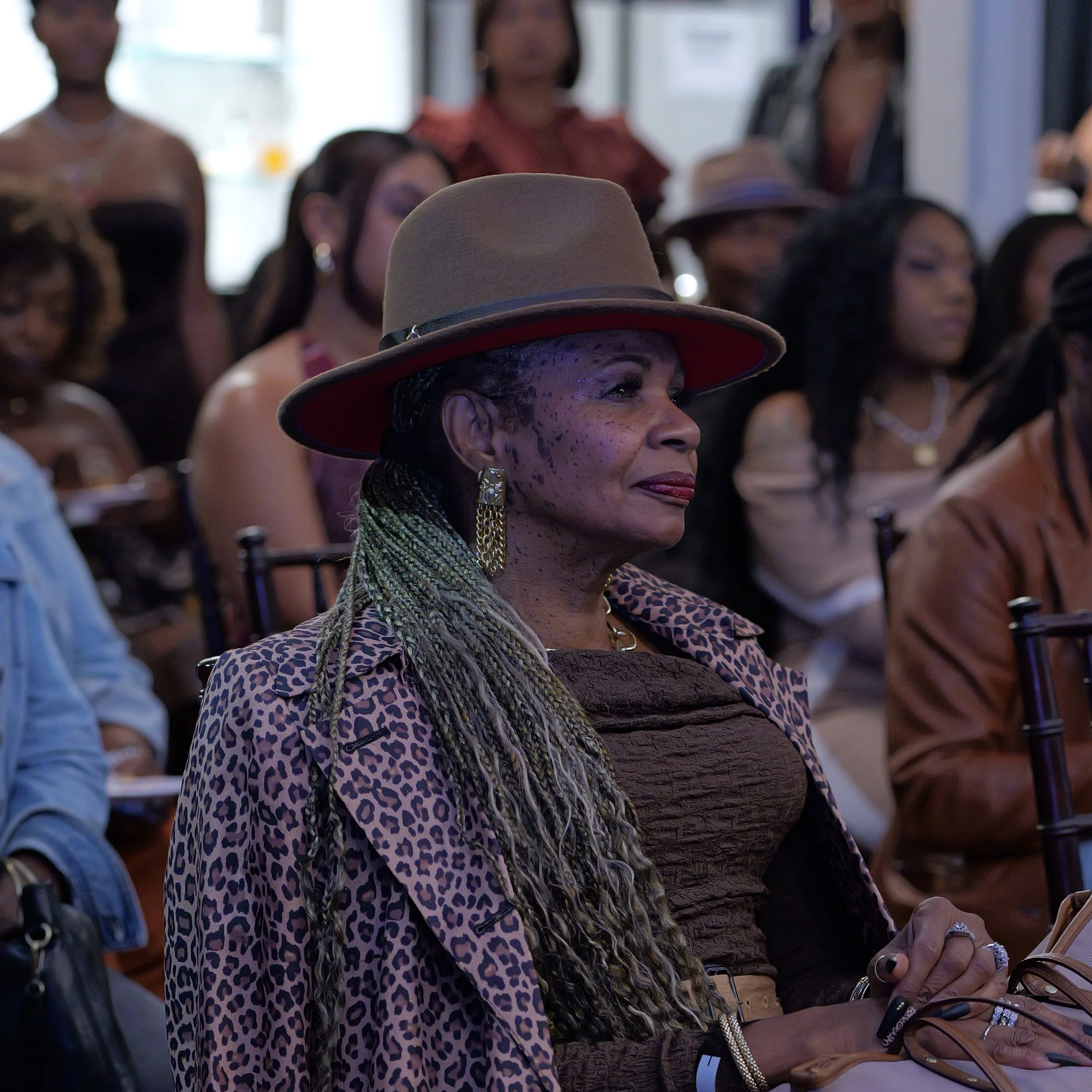An elderly woman with long braided hair, wearing a leopard print jacket, a brown top, a wide-brimmed hat, and gold earrings, attending an indoor event among other seated women.