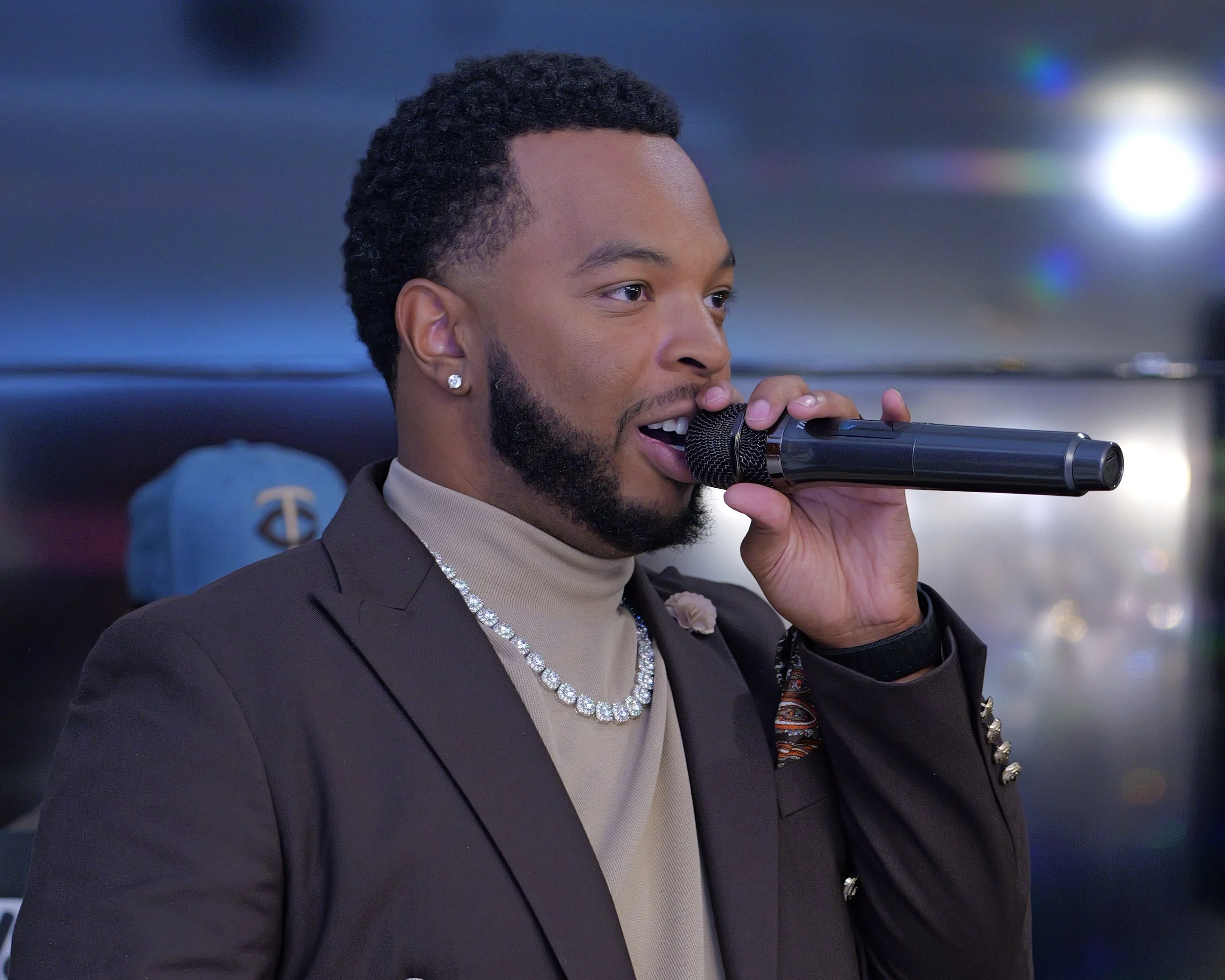 A man singing into a black microphone, dressed in a dark blazer, beige turtleneck, and wearing jewelry, with a woman and a hat in the background.