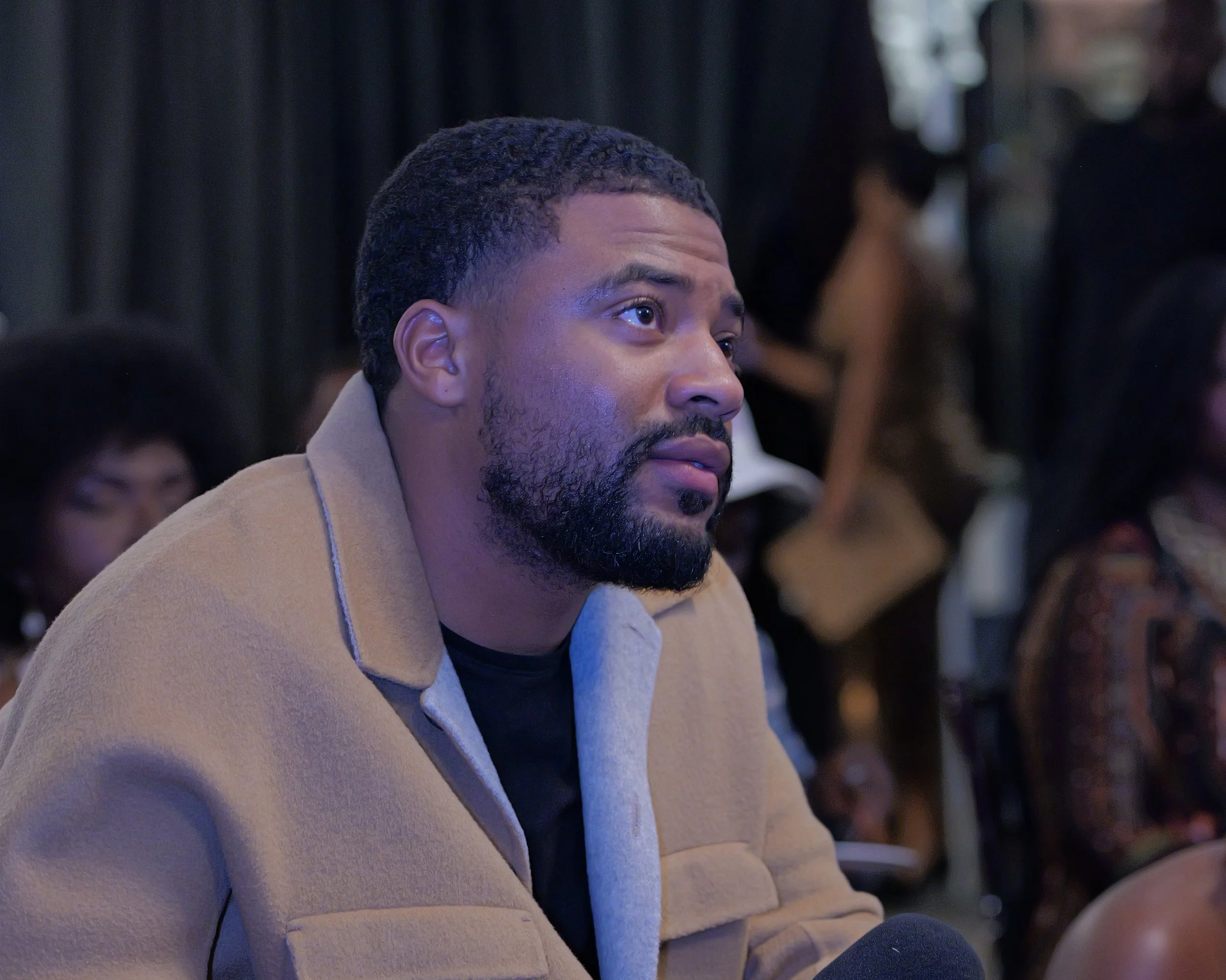 A man with a beard and short curly hair sitting attentively at an indoor event, wearing a beige coat over a dark shirt, with a woman and others in the background.