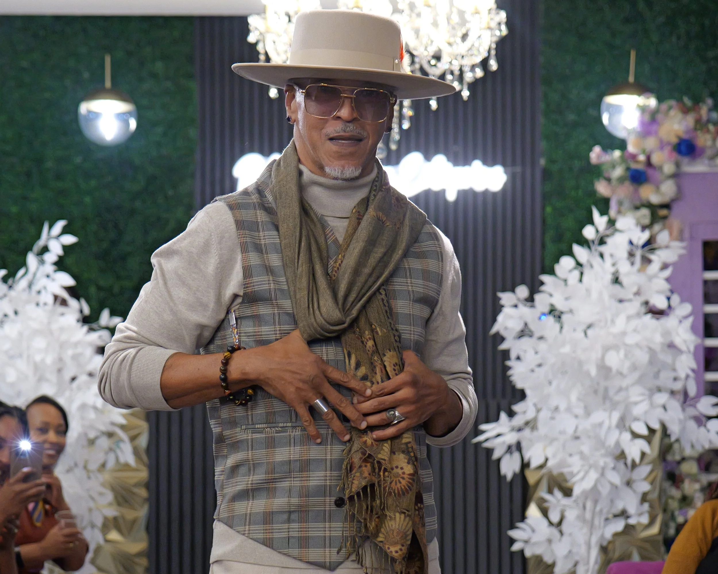 A man dressed in stylish clothing with a large hat, sunglasses, and scarf, standing indoors at a decorated event with white floral arrangements and a chandelier in the background.
