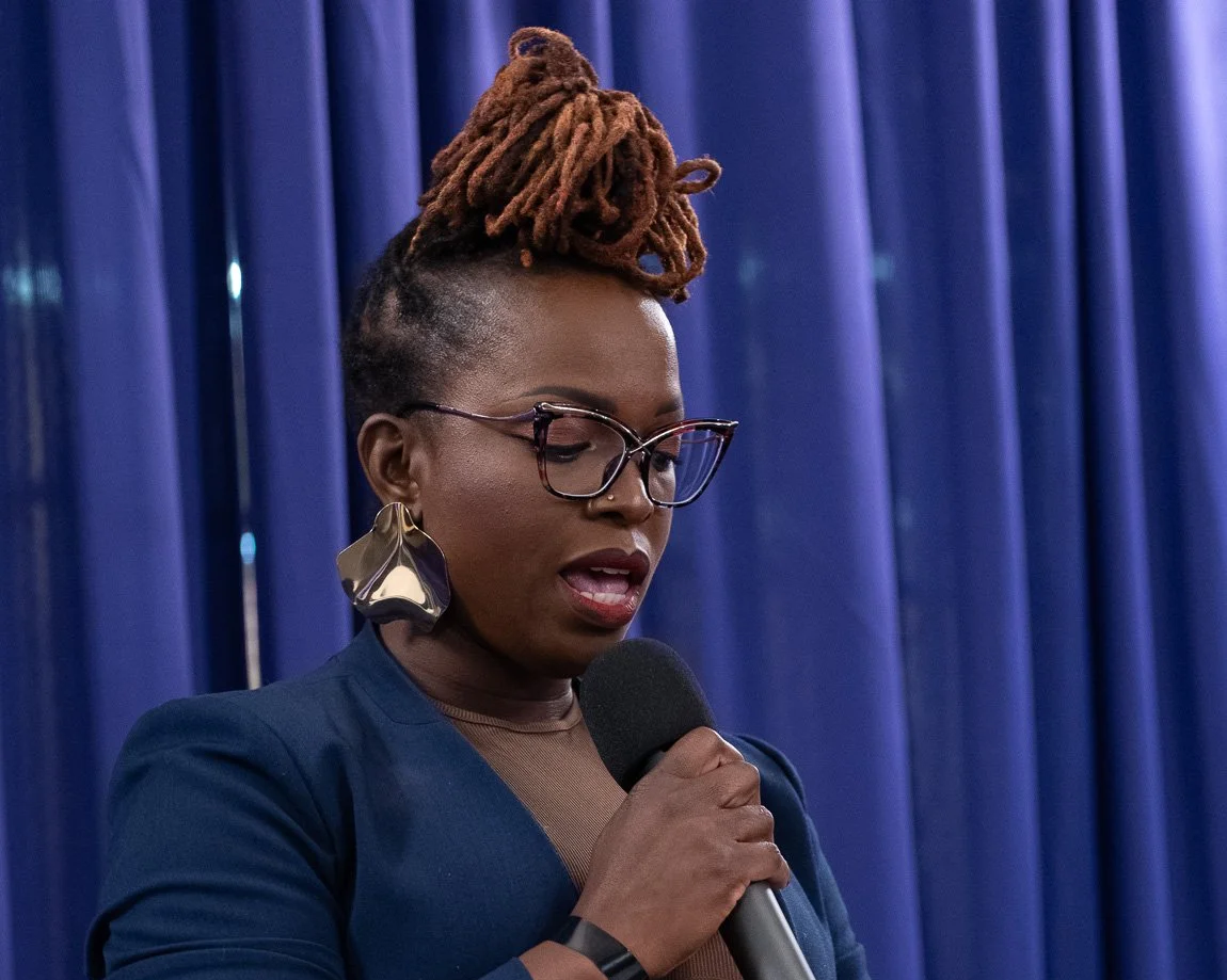 A woman with wrapped, dreadlocked hair styled in a high bun, wearing glasses, large earrings, a navy blazer, and holding a microphone, speaking in front of blue curtains.