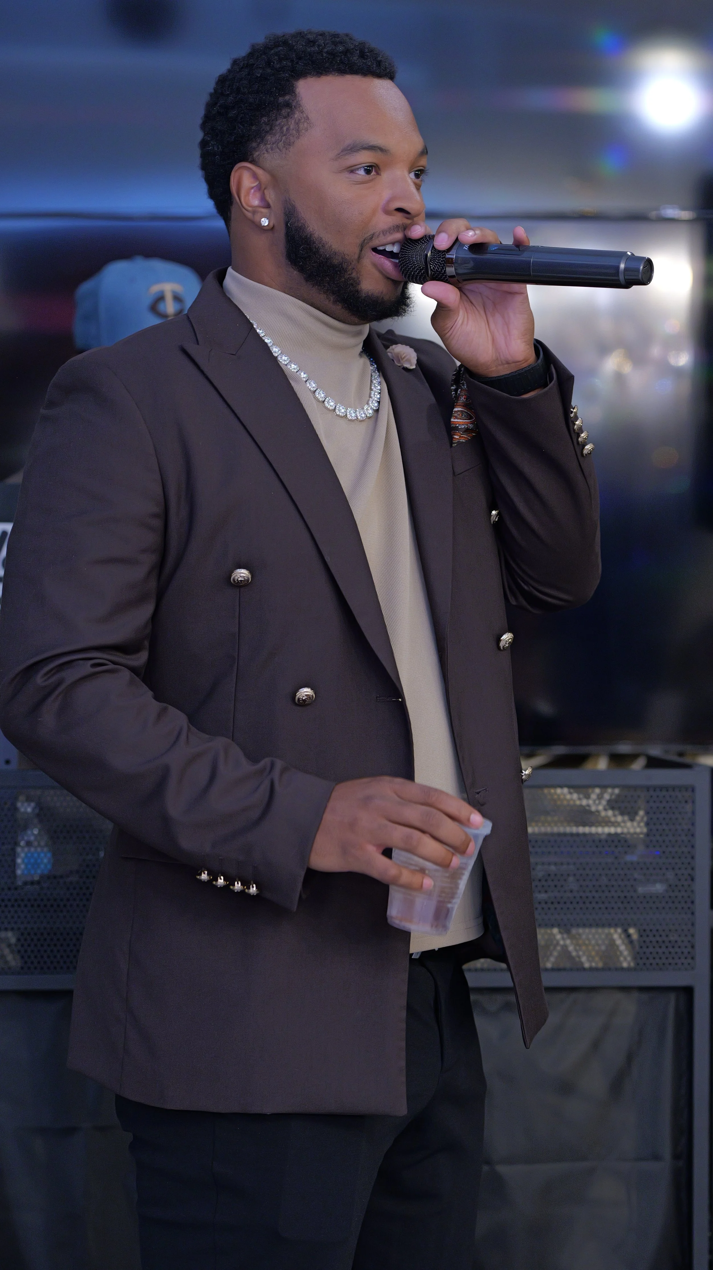 Man with a beard wearing a turtleneck, suit jacket, and diamond necklace, holding a microphone and a clear plastic cup, speaking in front of a blurred background with some lights.