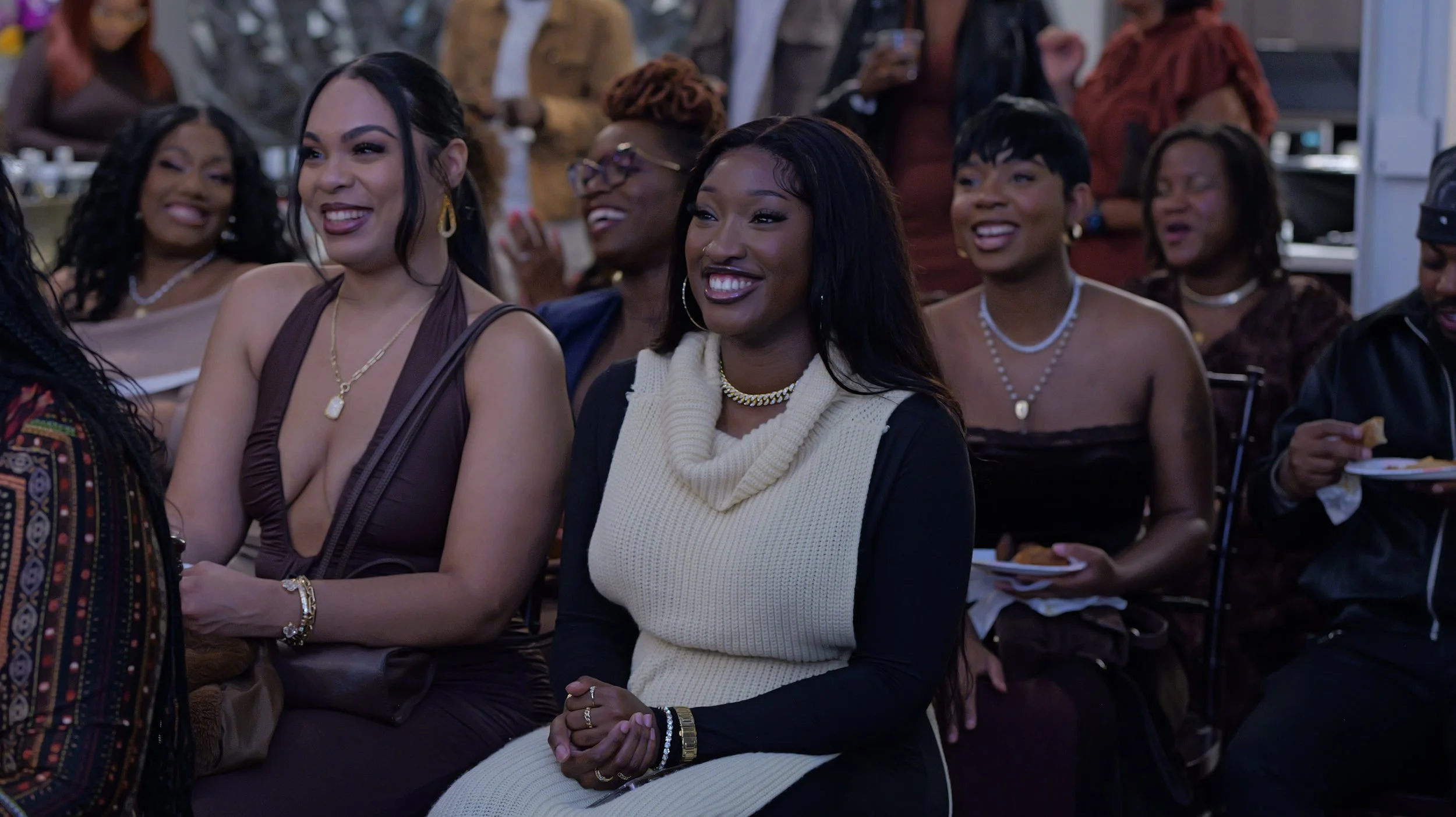 Group of women sitting and smiling at an event, with some holding plates of food, in a festive indoor setting.