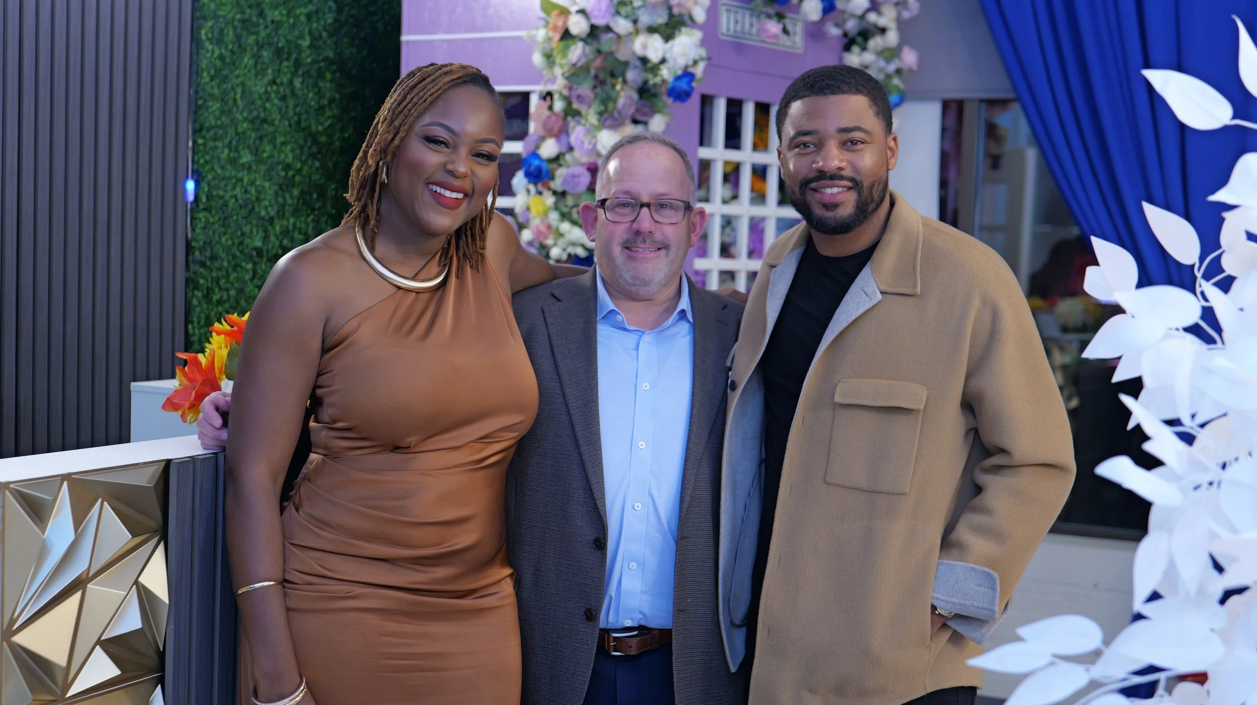 Three people standing together at an indoor event, smiling at the camera. A woman wearing a shiny bronze dress, a man in a blue shirt with a gray blazer, and a man in a black shirt with a tan jacket, with a floral and decorated background.