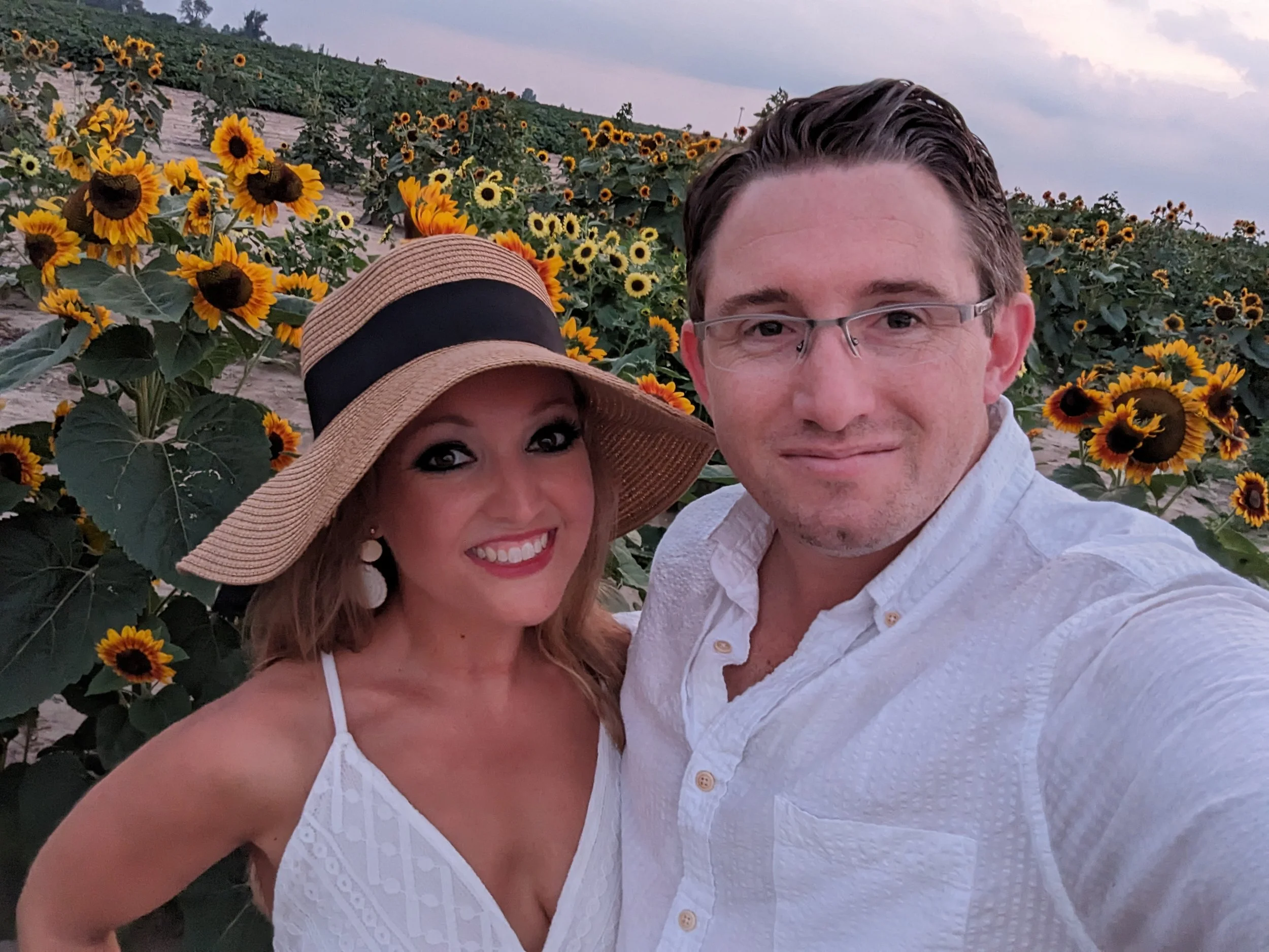 A couple taking a selfie in a sunflower field at sunset. The woman is wearing a wide-brimmed sunhat and white dress with spaghetti straps. The man is wearing a white button-up shirt and glasses.