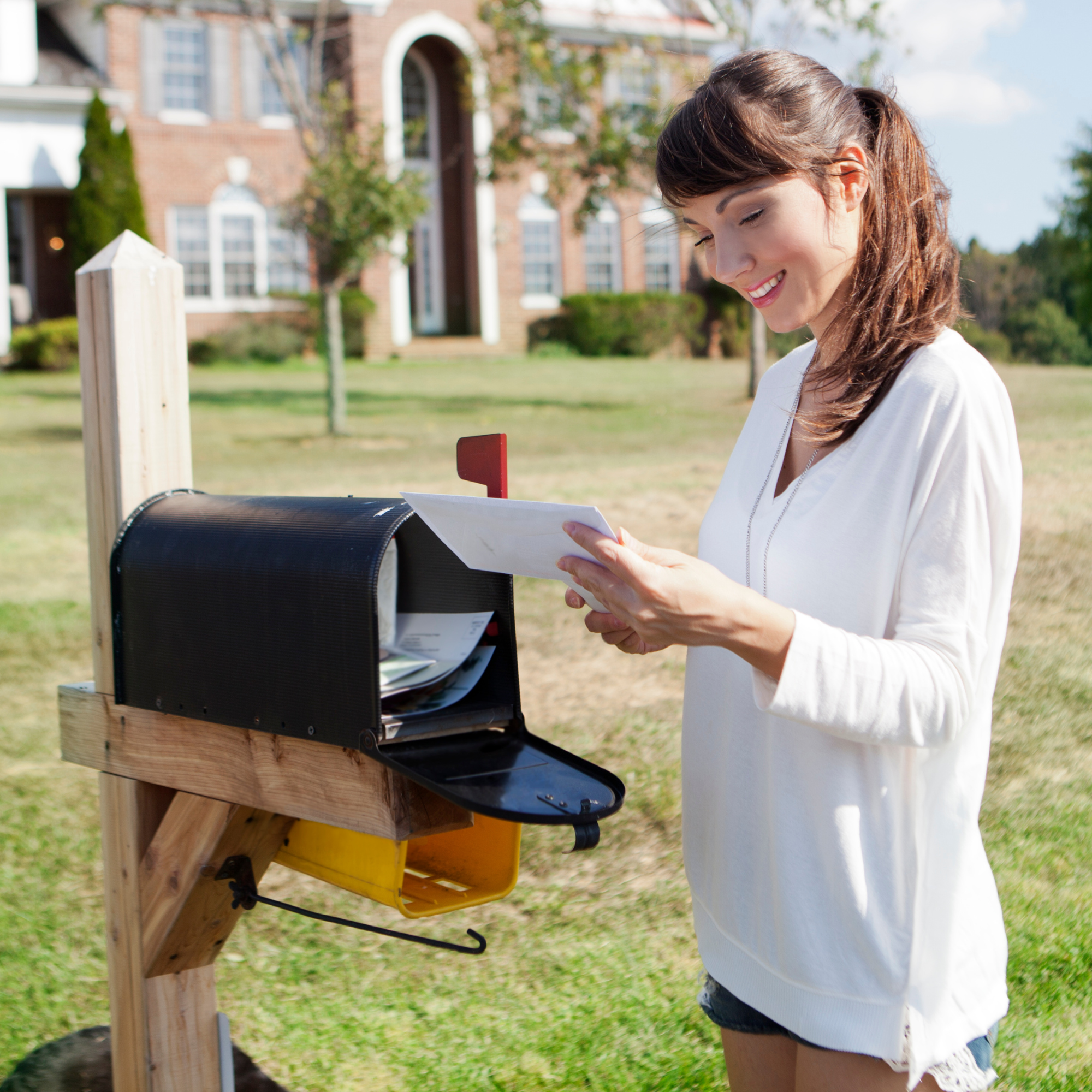 A woman in a white top and shorts dropping a letter into a mailbox on a wooden post outside near a house with green grass and trees in the background.