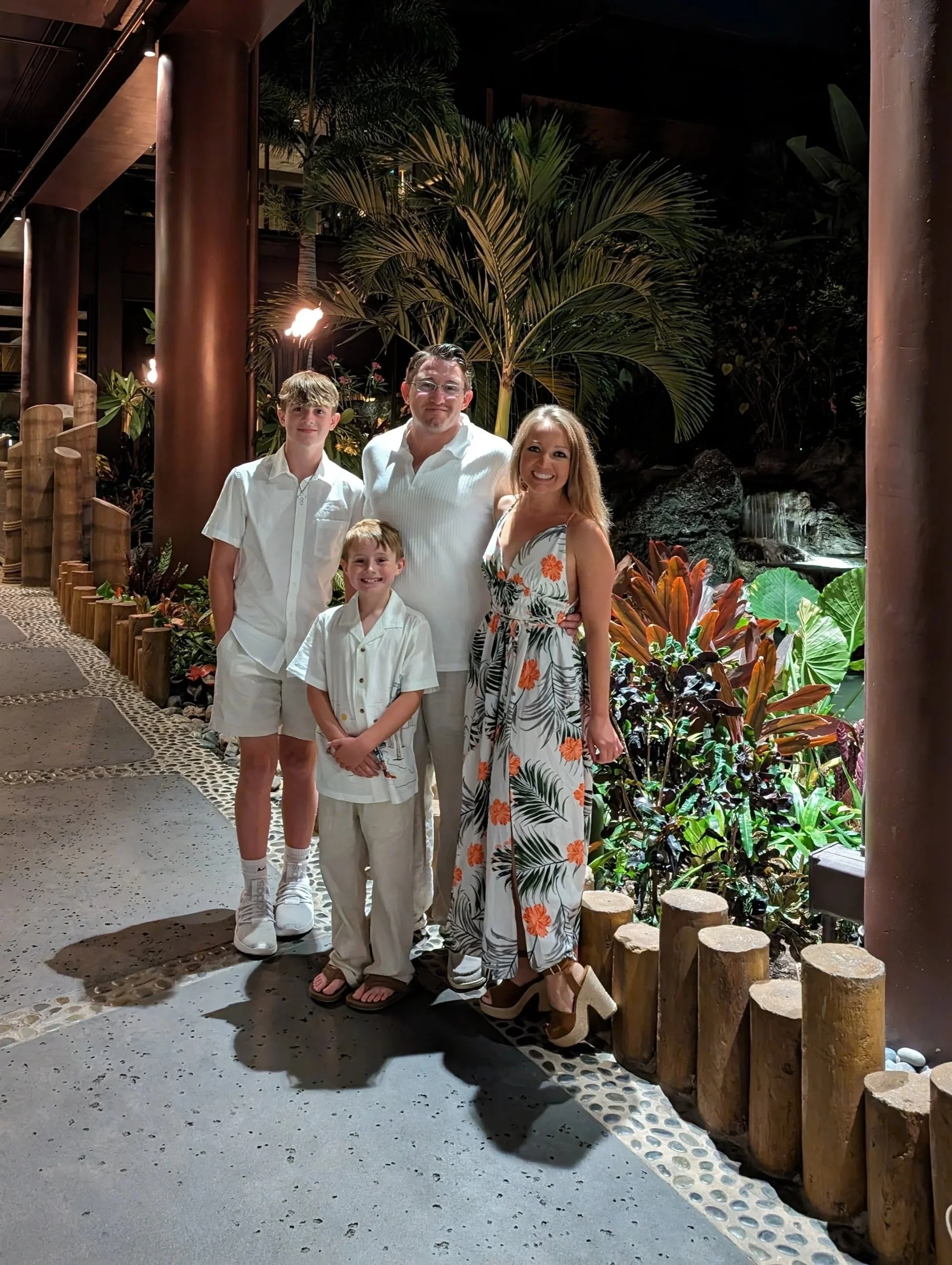 A family of four posing together outdoors at night in a tropical or garden setting with lush green plants and large leaves, illuminated by soft lighting.