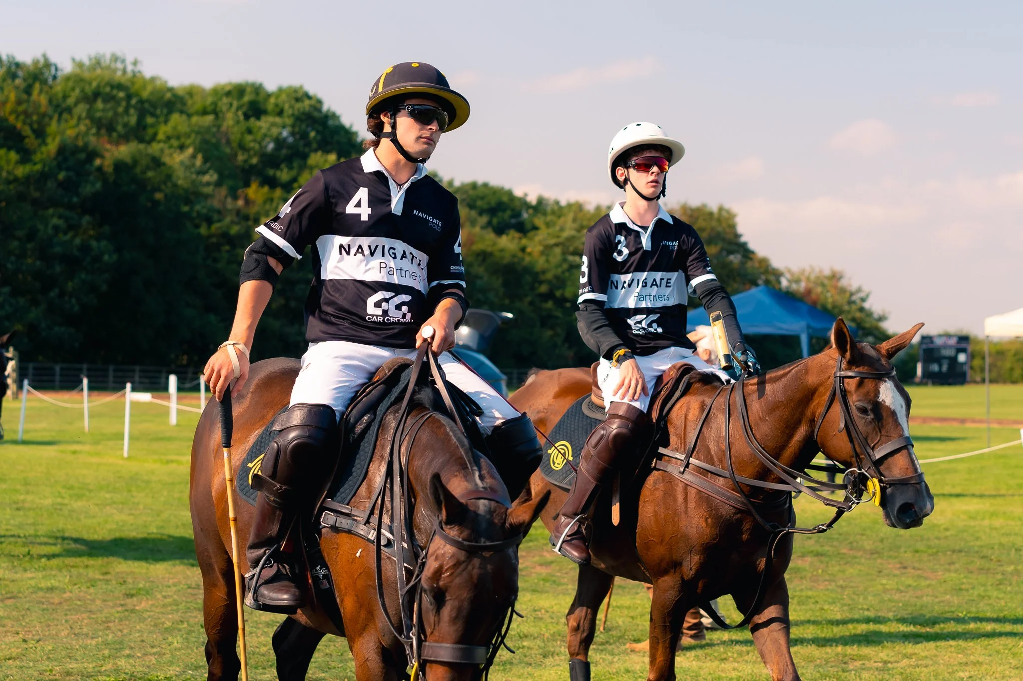 Two polo players riding horses on a grassy field, wearing helmets and black jerseys with white and blue accents.