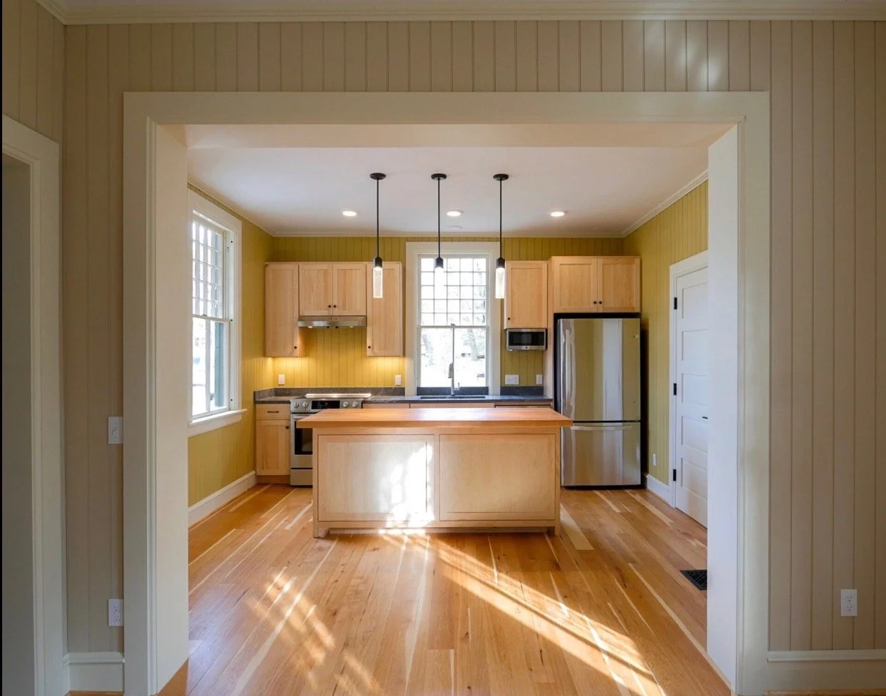 Modern kitchen with yellow walls, wooden cabinets, stainless steel appliances, a central island, hardwood flooring, and pendant lighting.