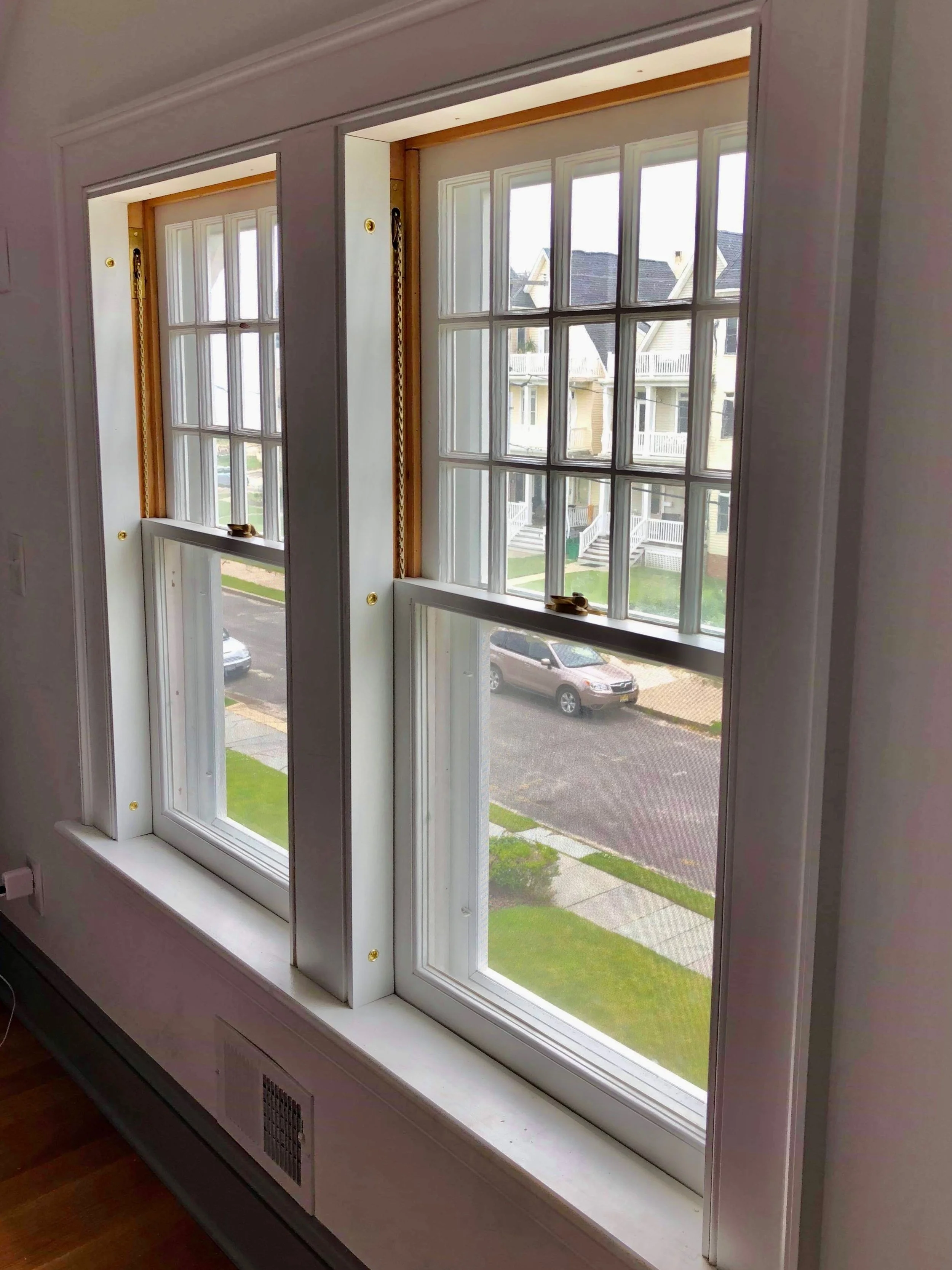 Interior view of a double window with white frames and divided panes in a house, overlooking a residential street with parked cars and front lawns.
