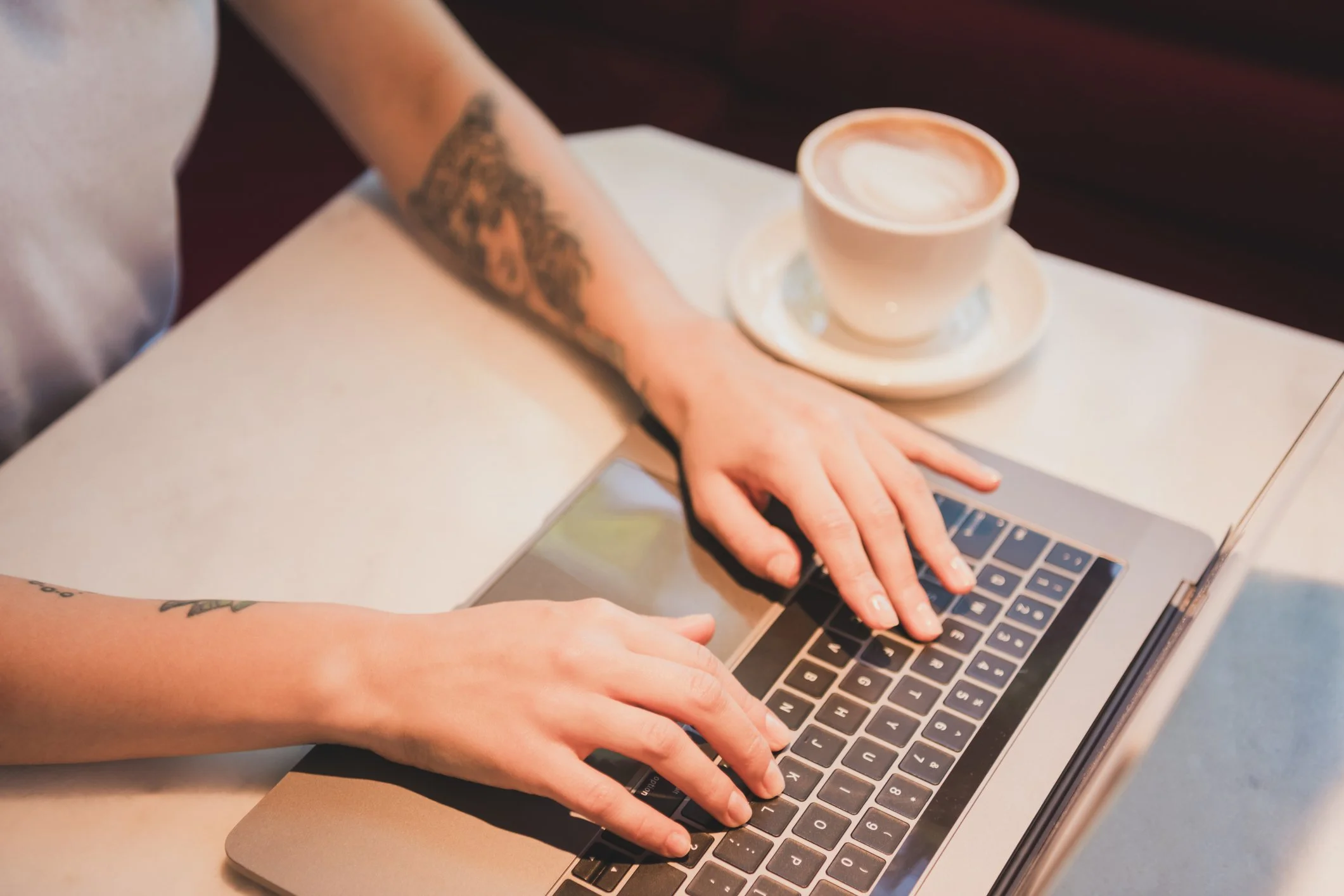 Person with tattoos on arms typing on a laptop with a cup of coffee nearby.