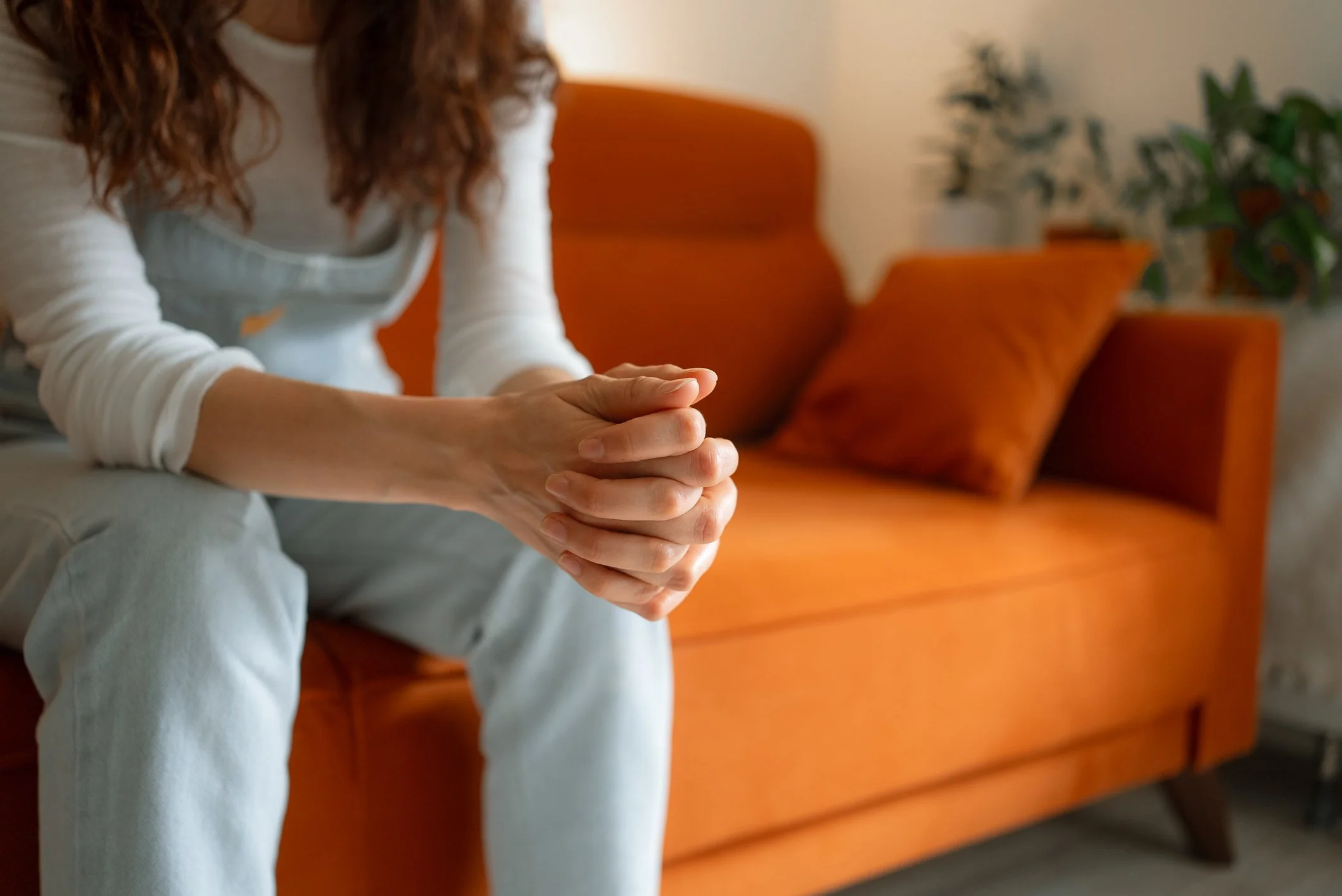 A woman with curly hair sitting on an orange sofa with her hands clasped together, in a room with a plant in the background.