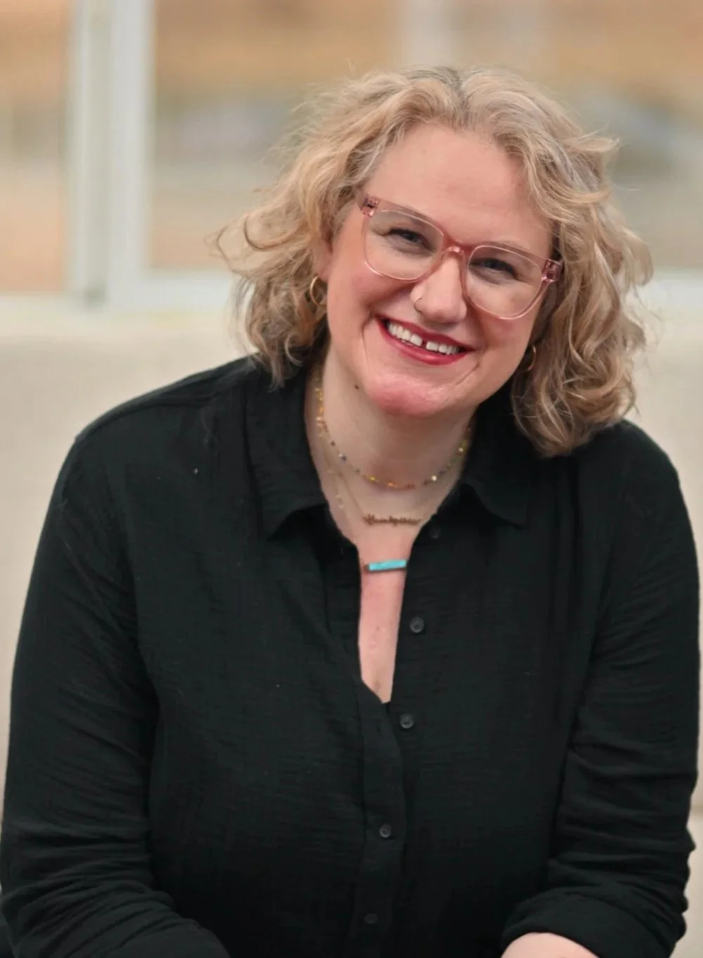 Smile of a woman with curly blonde hair wearing glasses and a black top, sitting indoors with a window in the background.