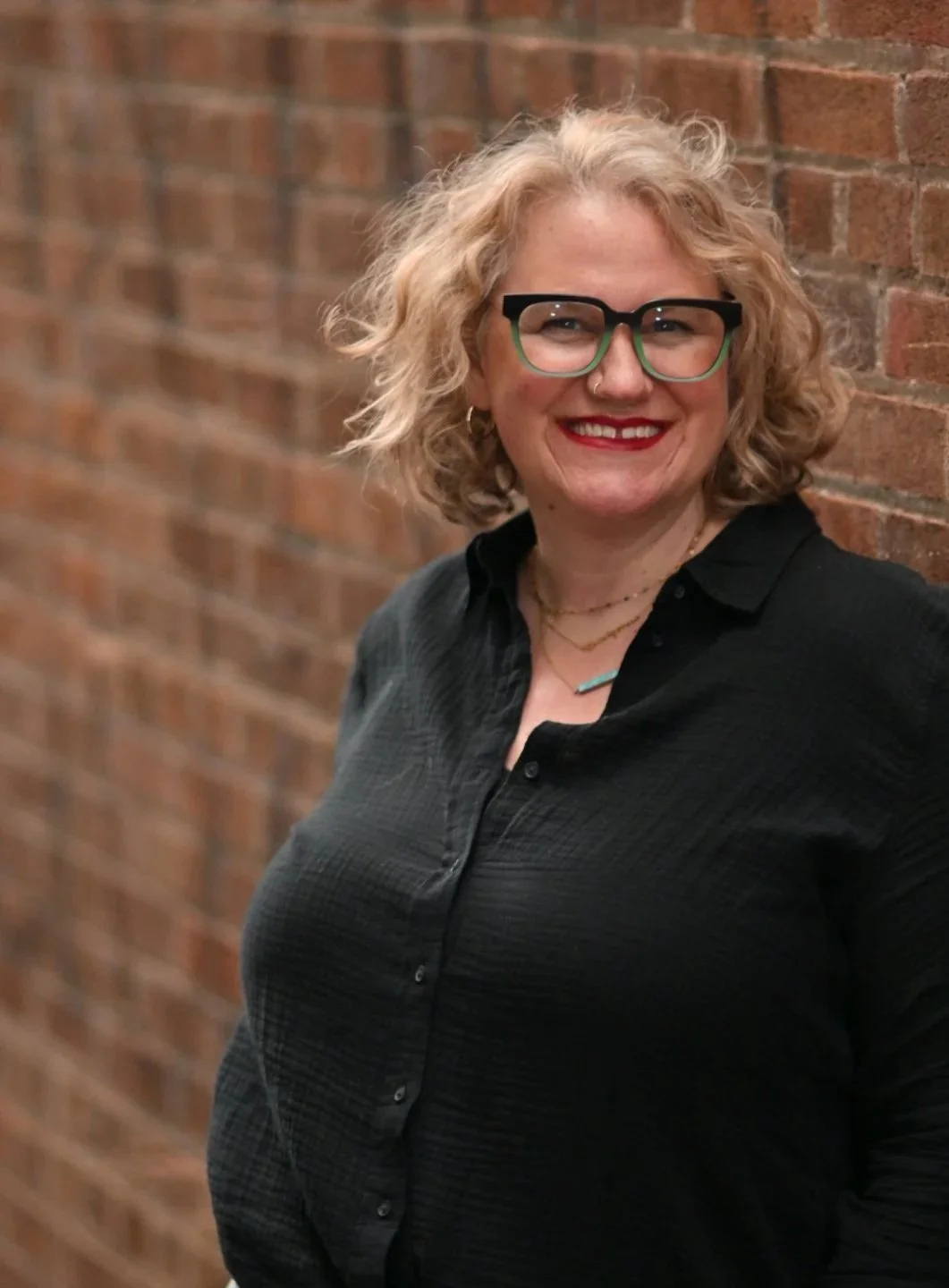 A woman with curly blonde hair, wearing glasses and a black button-up shirt, standing against a brick wall with a bright smile.