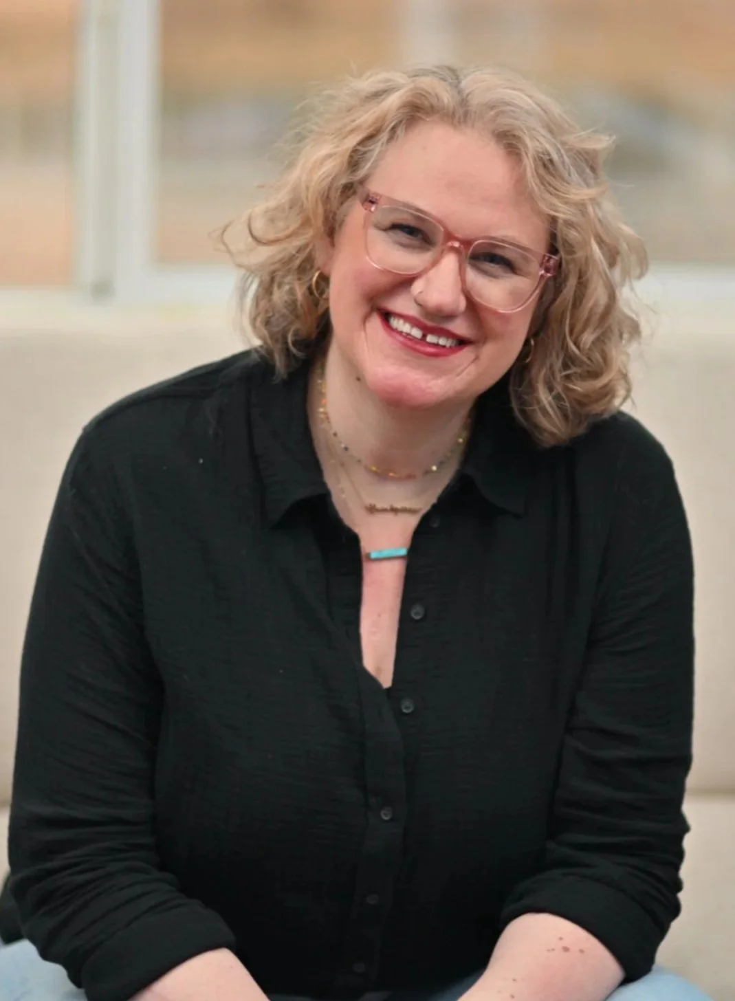 A woman with curly blonde hair, glasses, and red lipstick smiling, wearing a black shirt with rolled-up sleeves, jewelry, and sitting indoors near a large window.