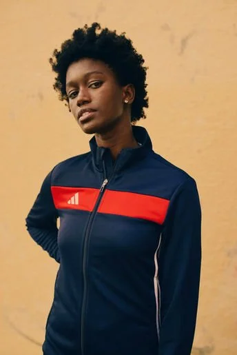 A young woman with short curly hair wearing a navy blue zip-up jacket with red and white stripes, standing against a yellowish wall.