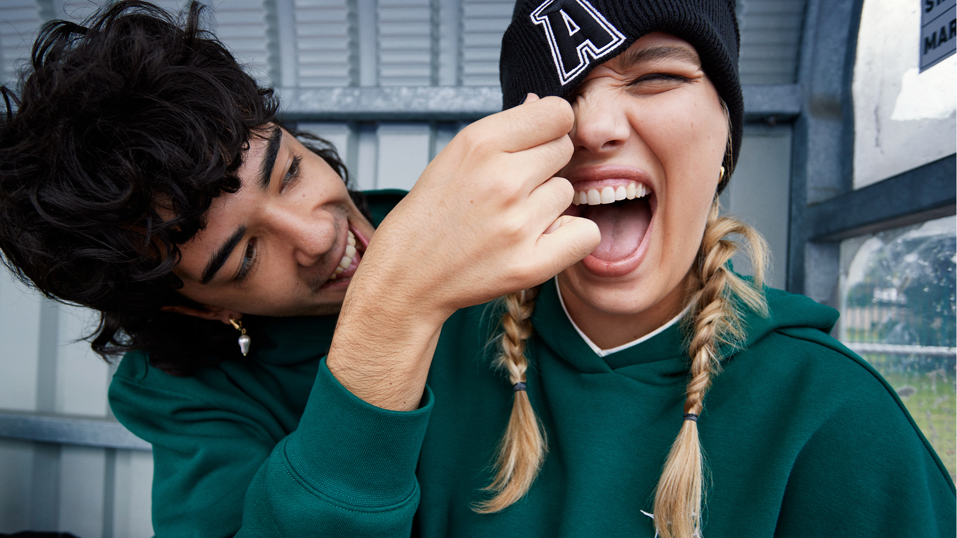 A man playfully pinching the nose of a smiling woman with blonde braids, wearing a black beanie and green hoodie, while leaning close to her. They are outdoors near a building with metal siding and an airplane window.