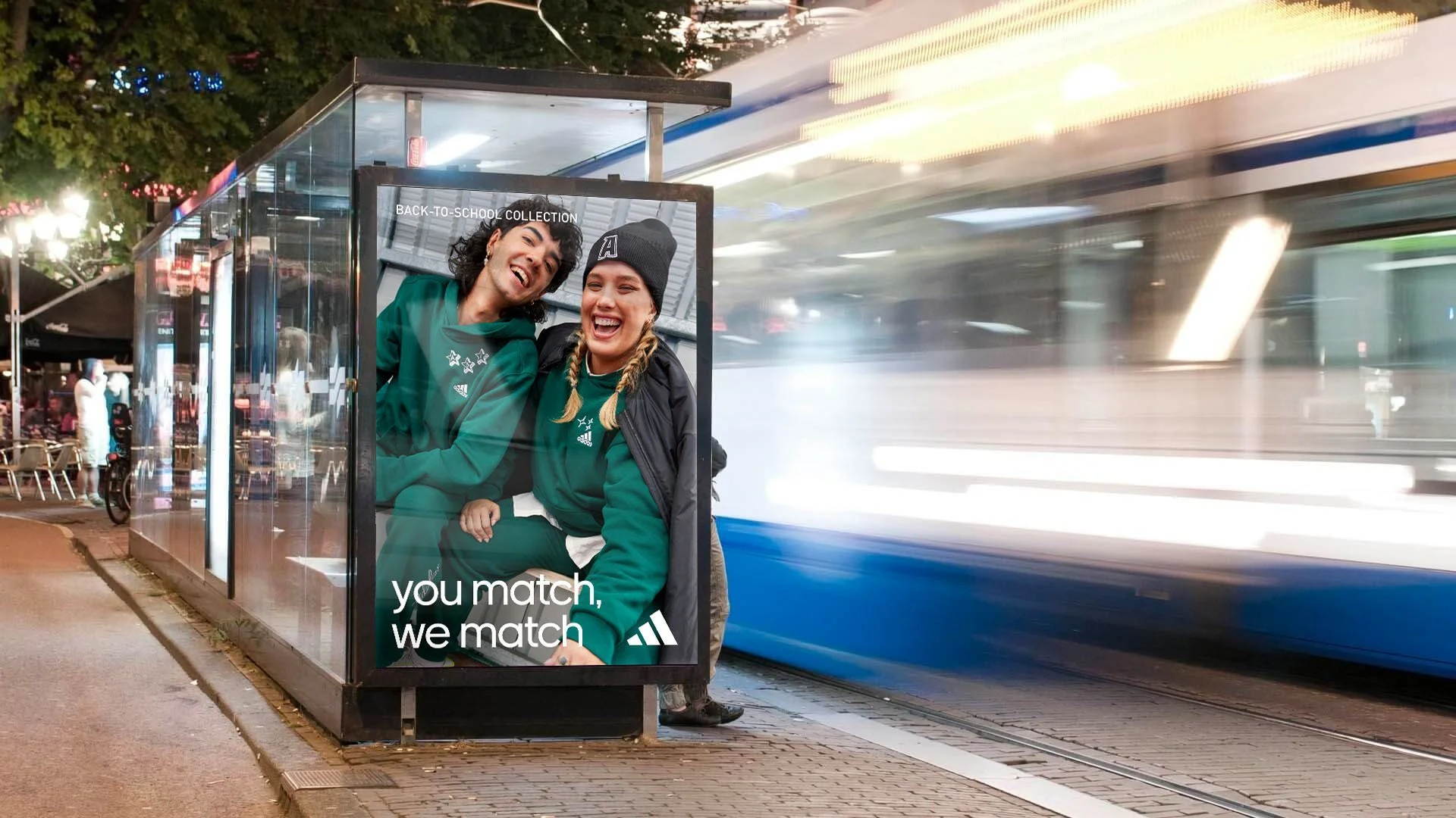 Two women sitting outside a bus shelter, wearing teal athletic hoodies, smiling and laughing, with a blurred city street and a passing tram in the background.
