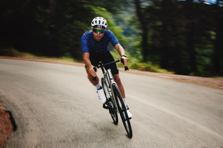 A man wearing a blue polka dot cycling jersey, black shorts, white socks, and a white helmet riding a road bike on a curved road surrounded by trees.