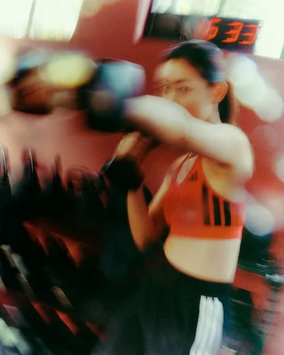 A woman in a sports bra and shorts boxing or training in a gym, with a digital clock in the background.