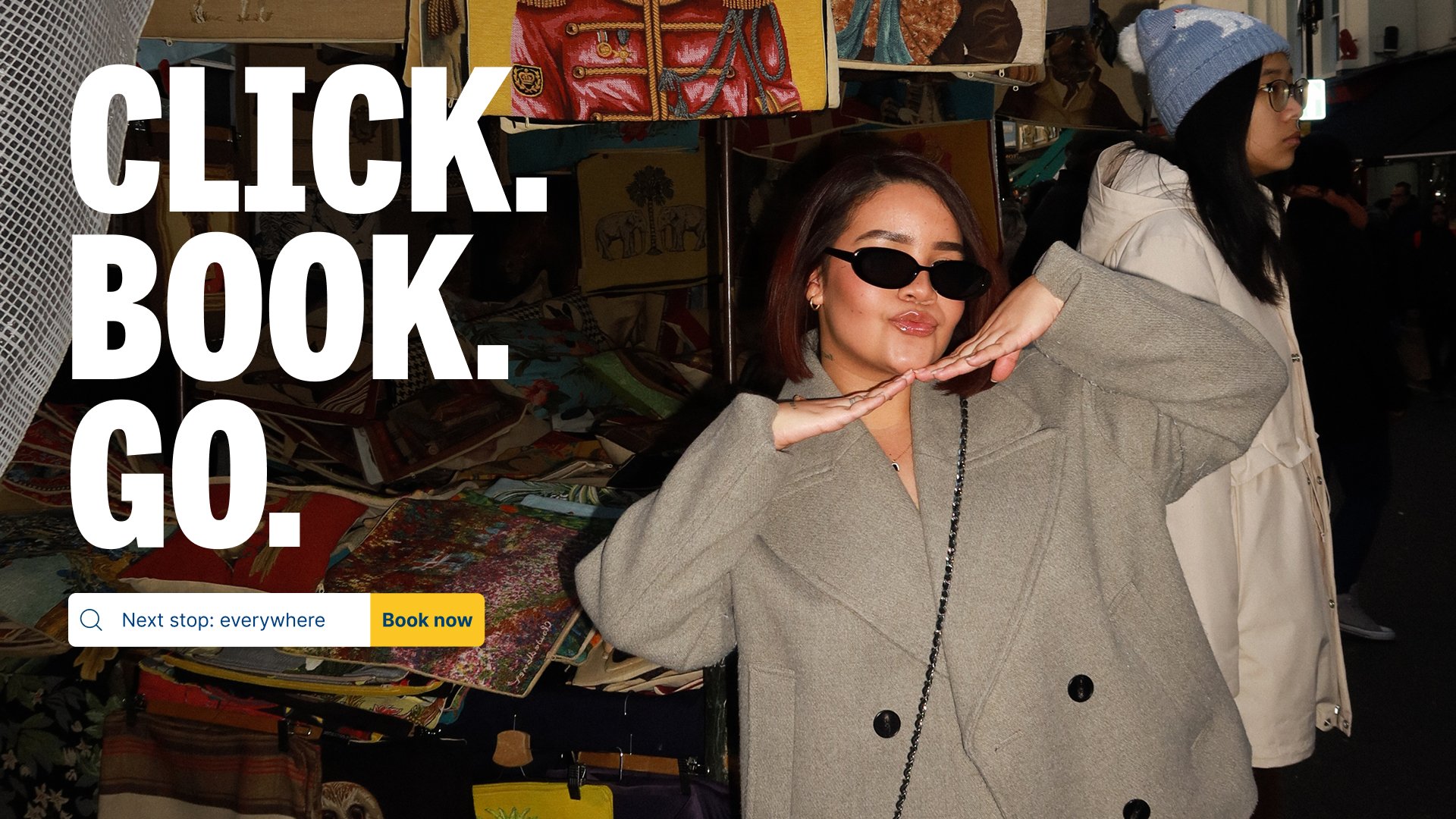 Woman in sunglasses and gray coat at a market stall with colorful fabric and artwork. Large white text overlay says, 'CLICK. BOOK. GO.' and a smaller button says 'Book now' with the phrase 'Next stop: everywhere.'