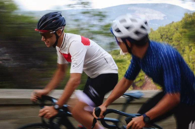 Two cyclists wearing helmets riding on a road surrounded by greenery and trees.