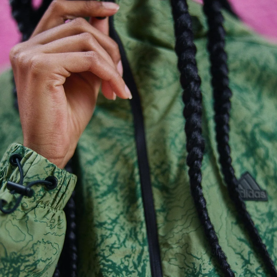 Close-up of a person's hand adjusting the drawstring of a green Adidas jacket with black braided hair visible in the background.
