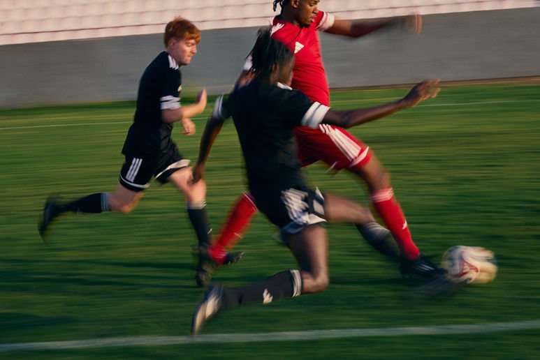 Three young soccer players in black and red uniforms chasing a soccer ball on a grass field during a game.