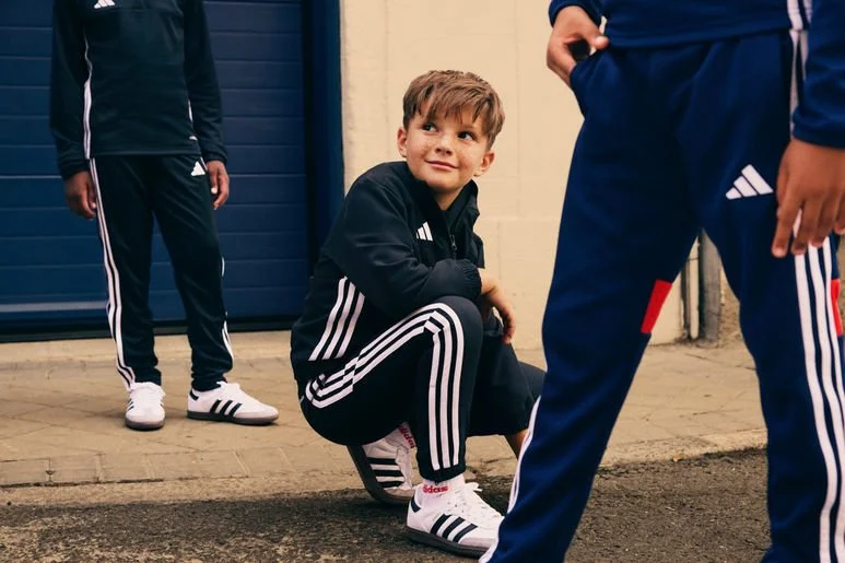 Young boy in black Adidas tracksuit squatting on sidewalk, smiling, with two other people in athletic attire nearby outside a building.
