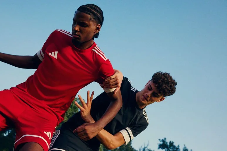 Two young men engaged in a soccer match, one in a red uniform and the other in black, outdoors against a clear blue sky.