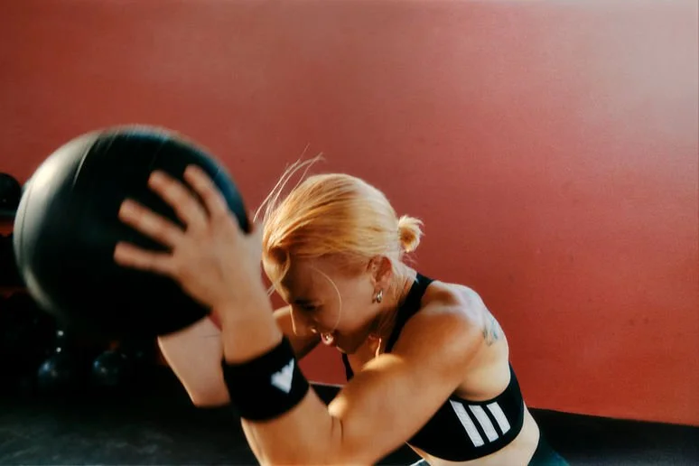 A woman with blond hair in a ponytail is exercising with a medicine ball in a gym, against a reddish wall.
