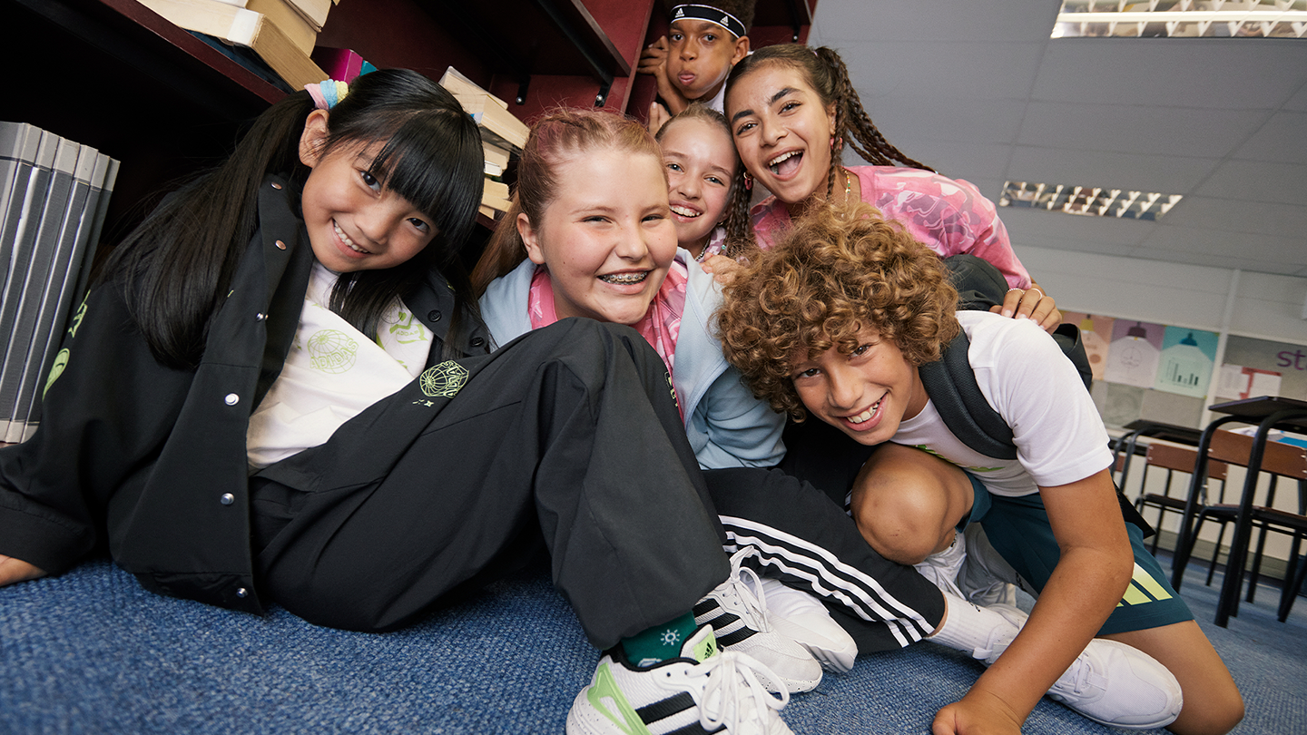 Group of six children smiling and playing together on the floor in a classroom, with bookshelves and desks in the background.