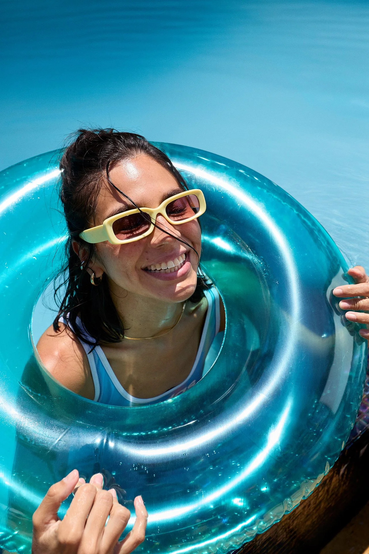 Smiling woman in sunglasses and a striped swimsuit, holding a blue inflatable pool float, smiling in a swimming pool under sunny sky.