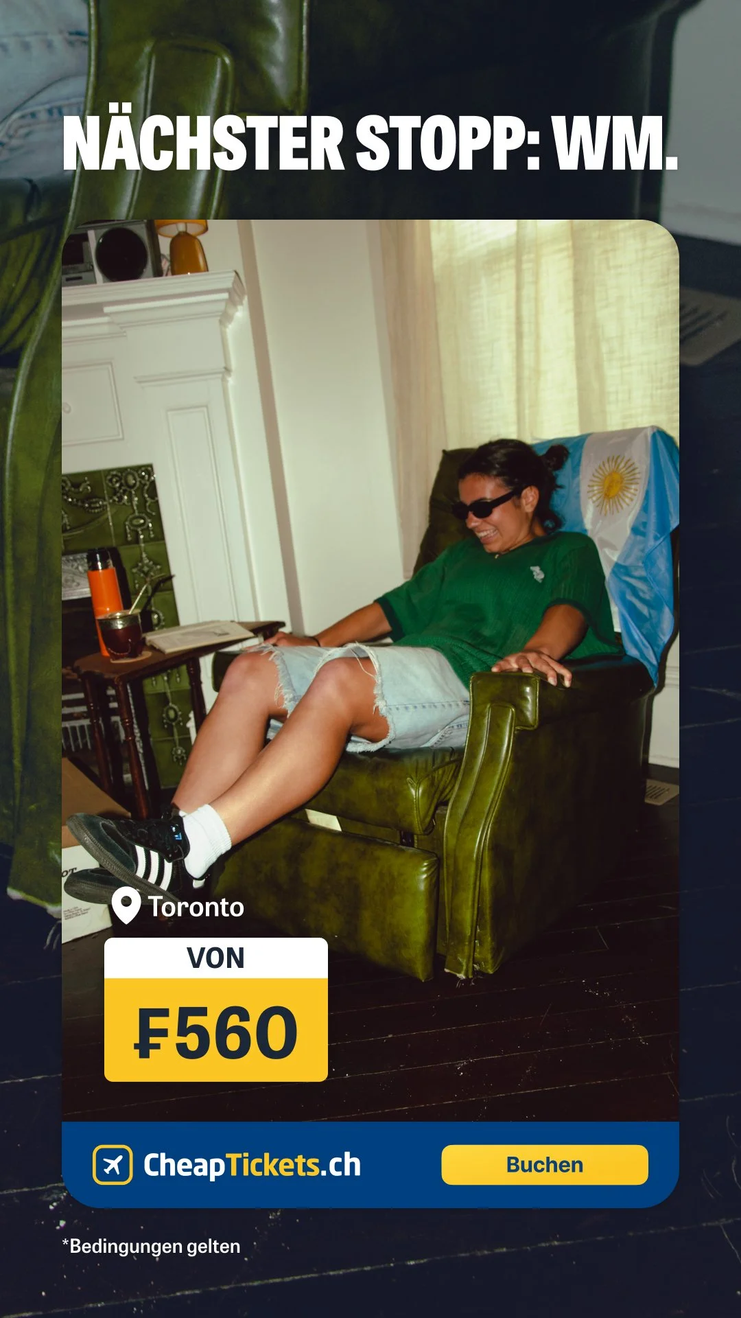 A person sitting in a green armchair with a Roman flag draped over the back of the chair. They are wearing sunglasses, a green t-shirt, and denim shorts while smiling. There is a side table with a drink and a book nearby, and a window with curtains i