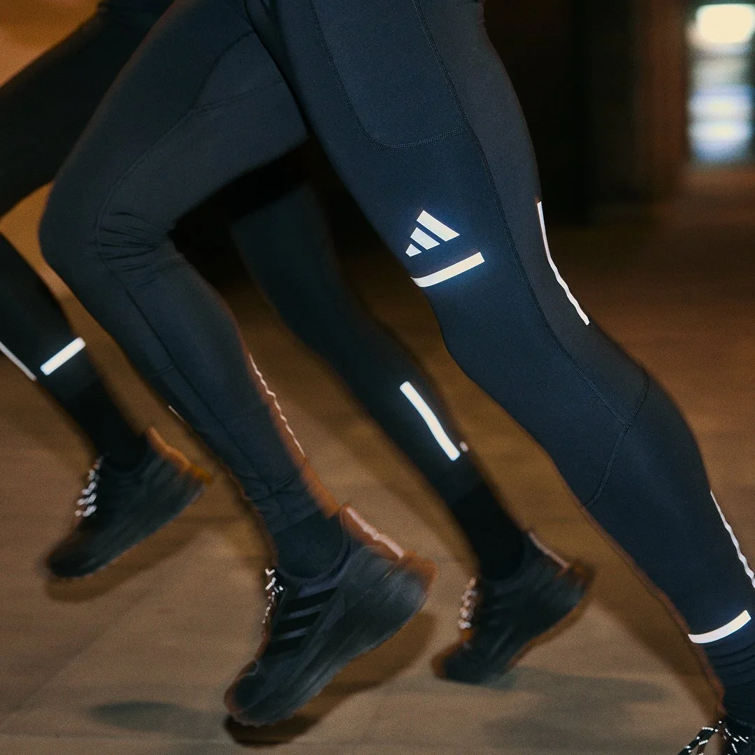 Close-up of people wearing black athletic leggings with reflective stripes and running shoes, performing a workout or dance move indoors.