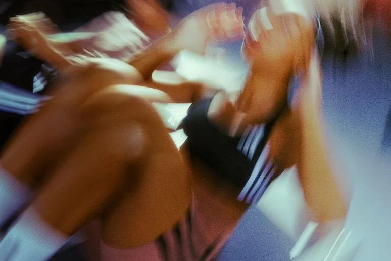 A person with long hair in a black sports bra and shorts sitting on a gym bench, lifting a barbell, with other people blurred in the background.
