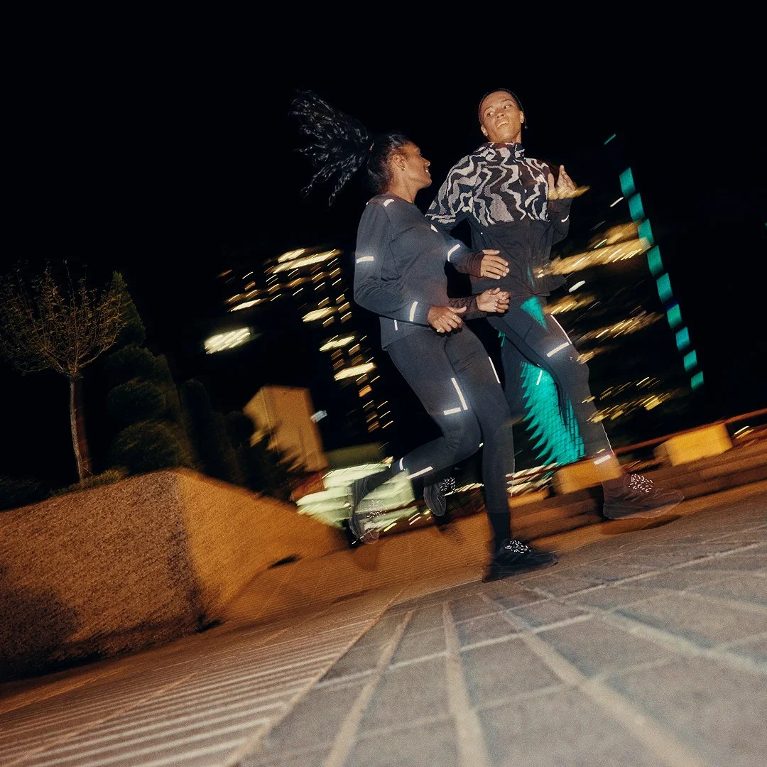 Two women running at night on a city street, illuminated by city lights and wearing reflective clothing.